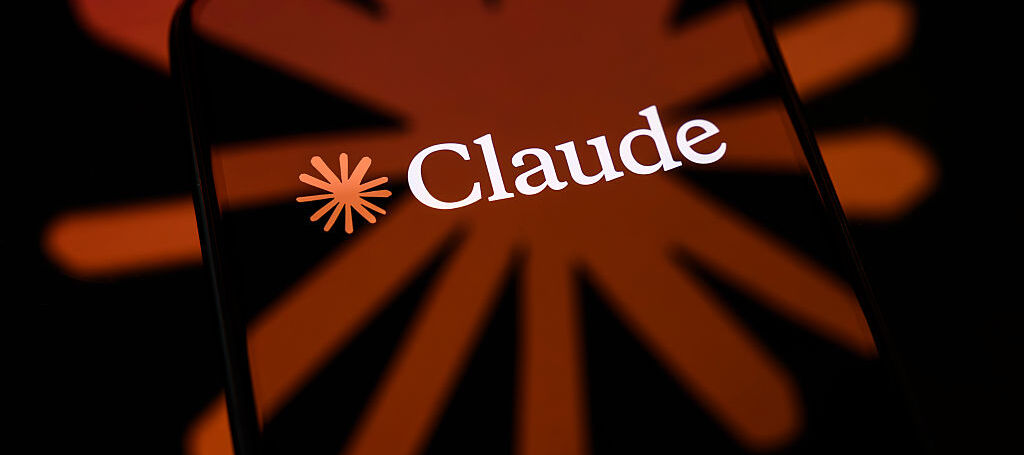 The Claude logo is displayed on a smartphone screen placed on a reflective surface on which the icon of the AI model is projected.