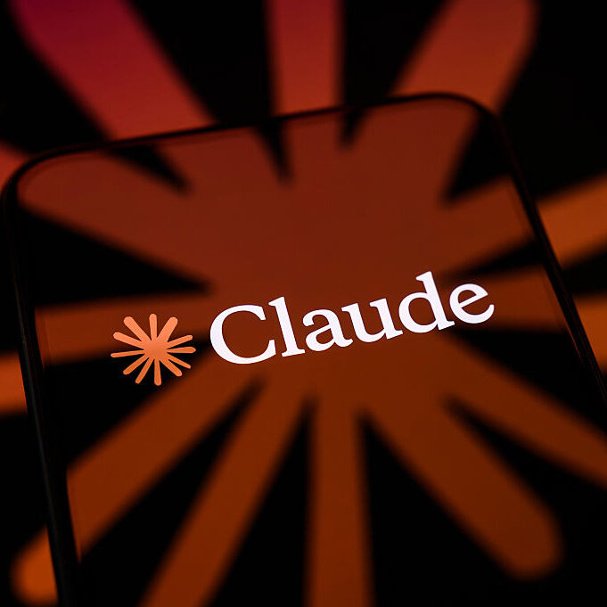 The Claude logo is displayed on a smartphone screen placed on a reflective surface on which the icon of the AI model is projected.
