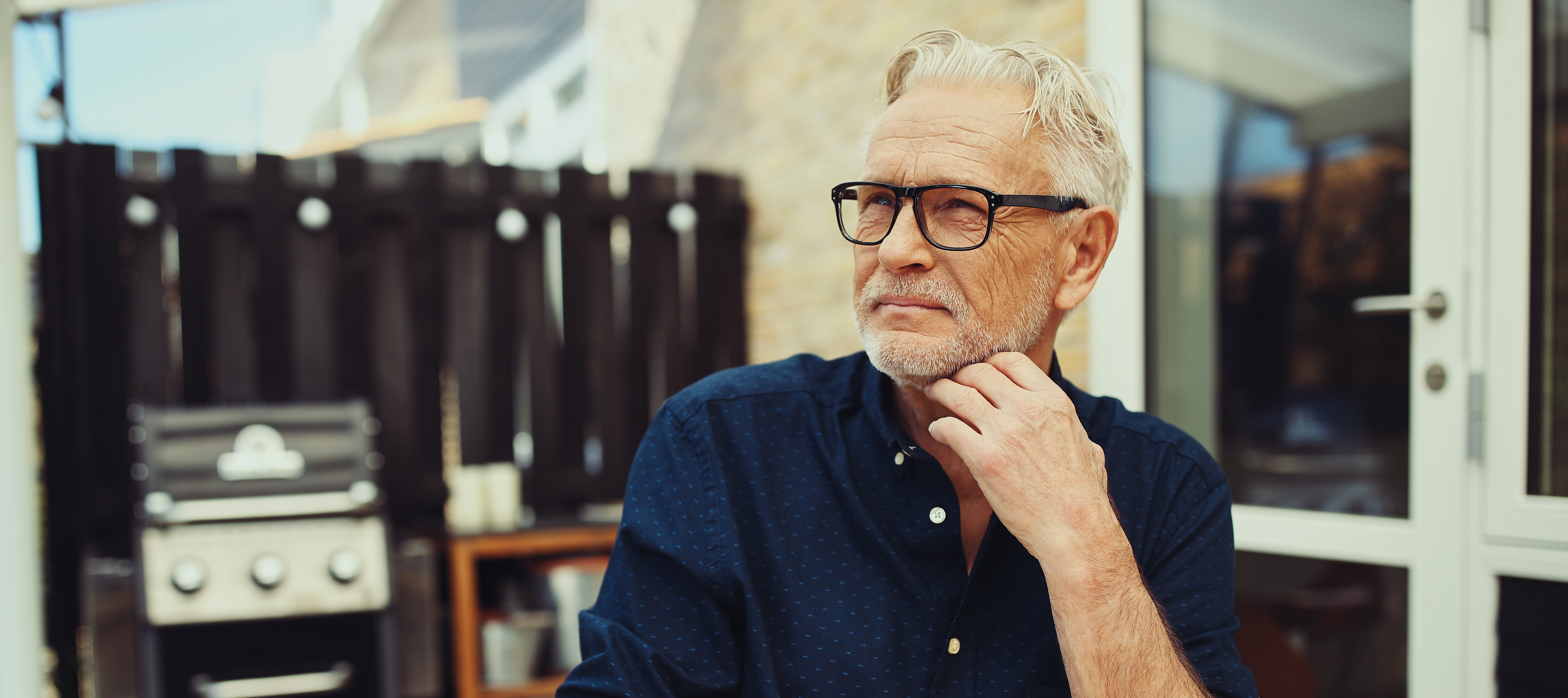A man with grey hair and glasses is sitting at a table outdoors reading in his backyard.