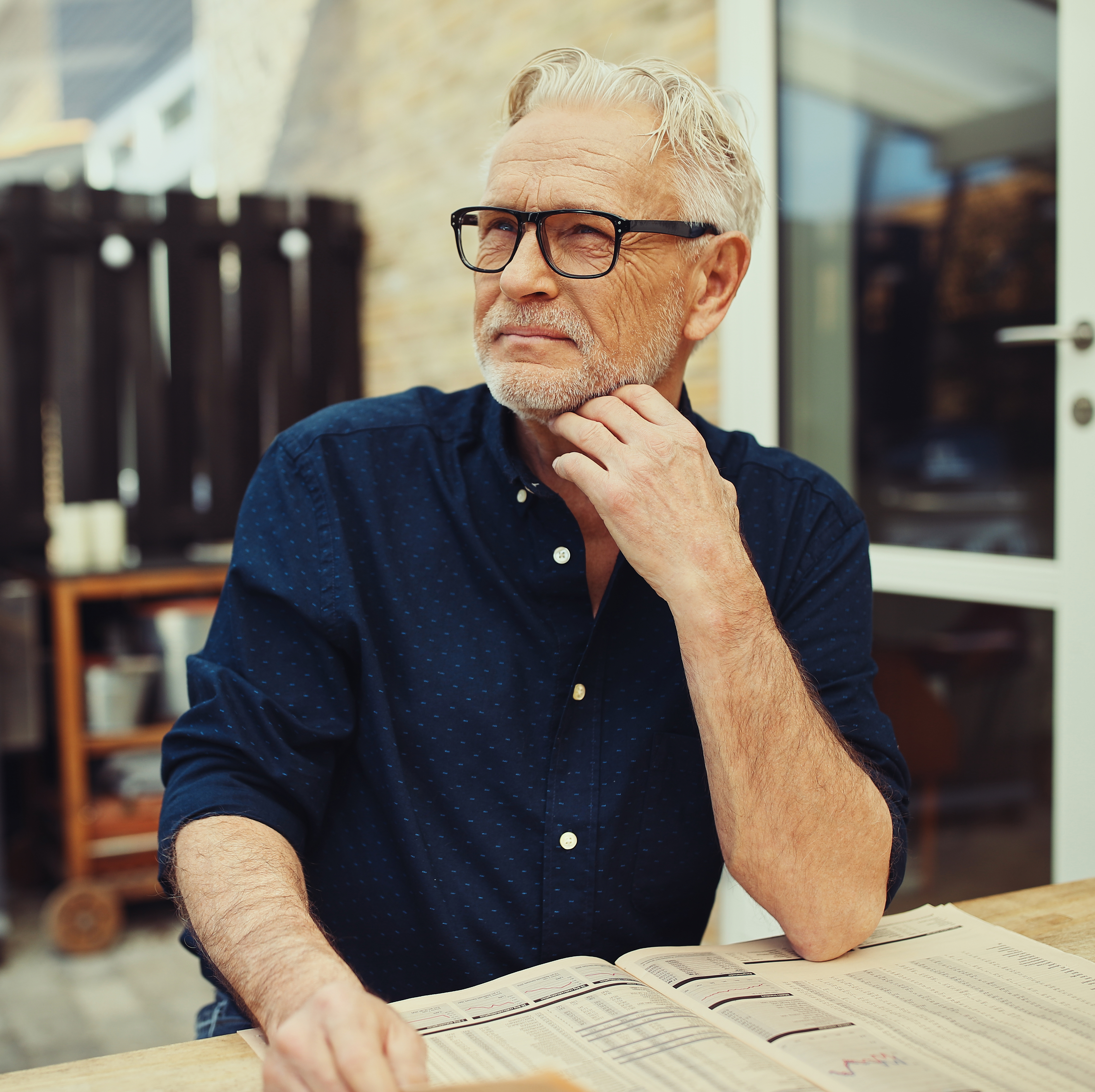 A man with grey hair and glasses is sitting at a table outdoors reading in his backyard.