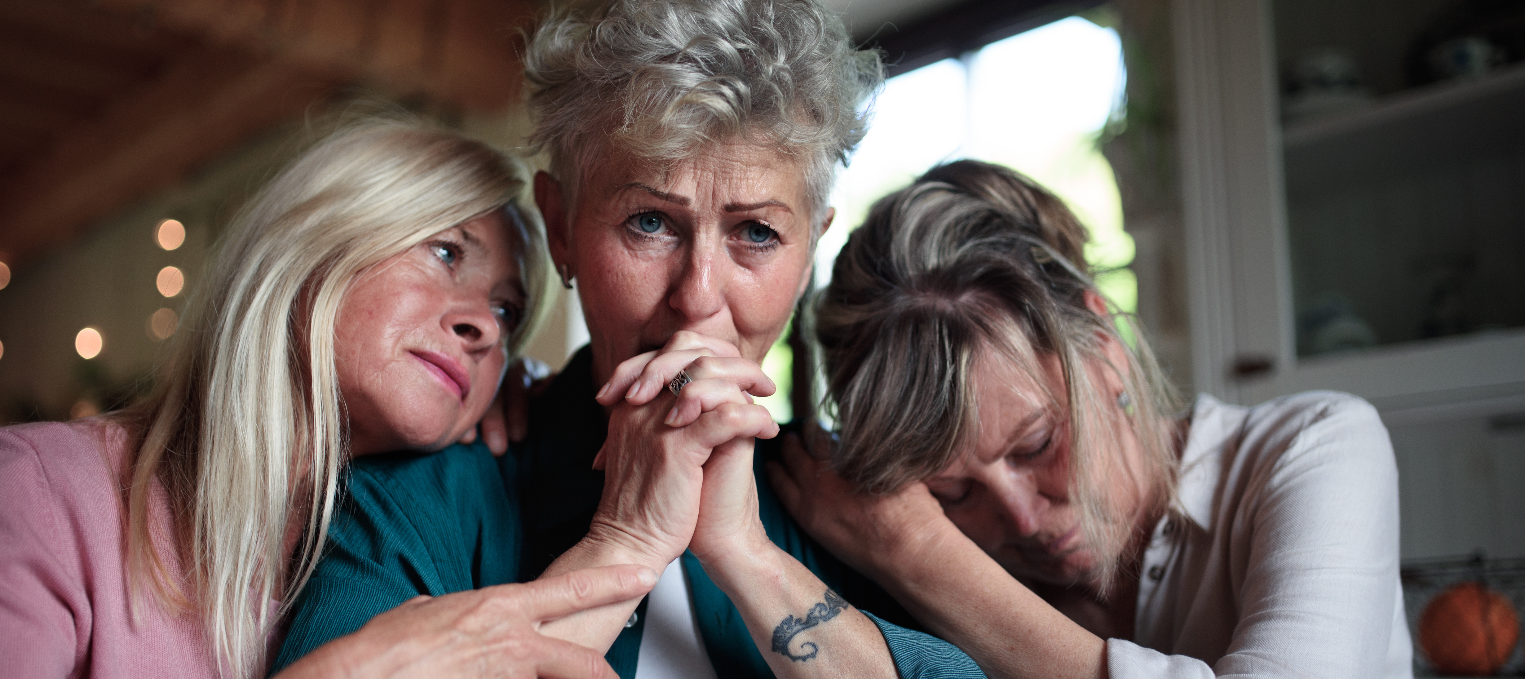 Three women gather indoors, sharing an emotional moment.