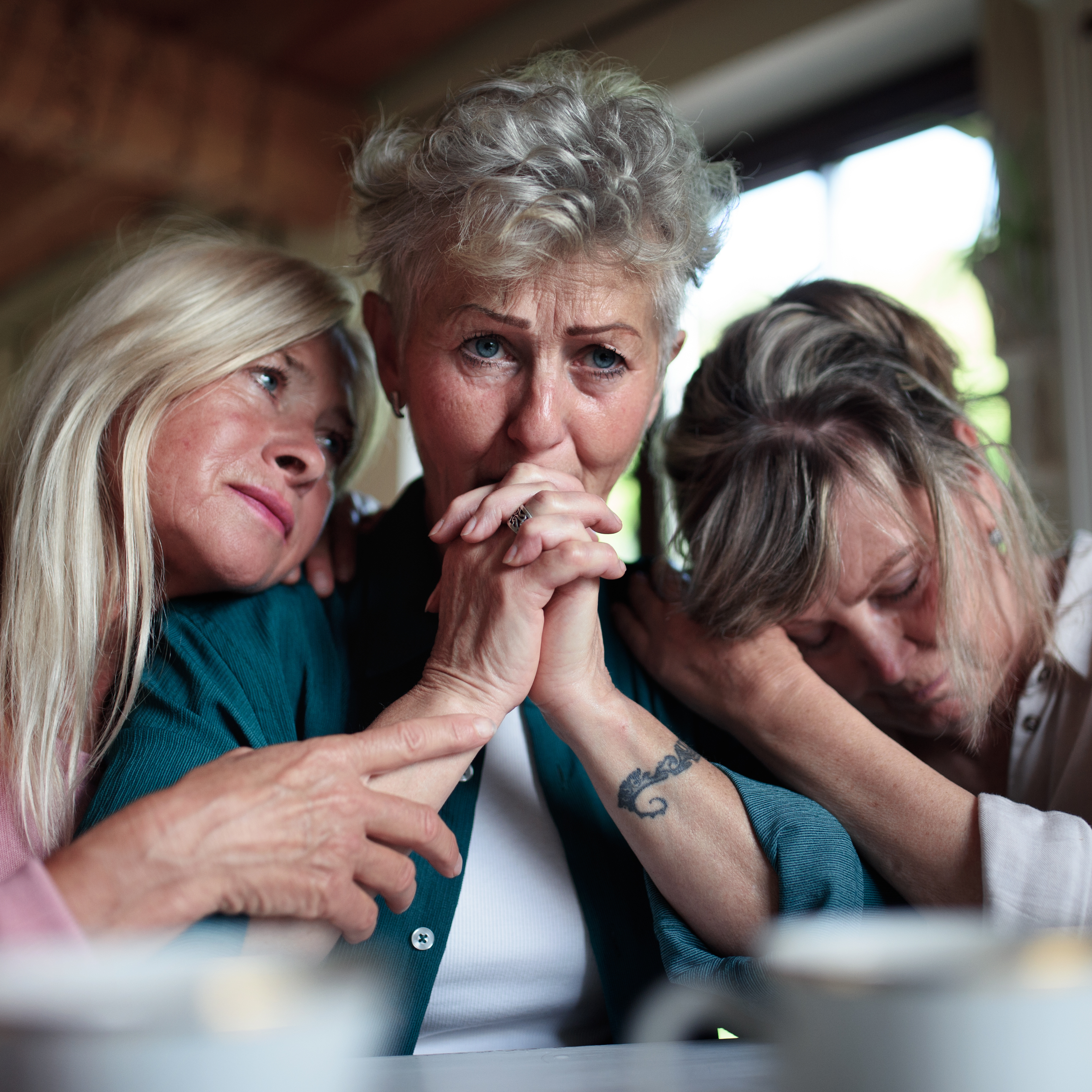 Three women gather indoors, sharing an emotional moment.