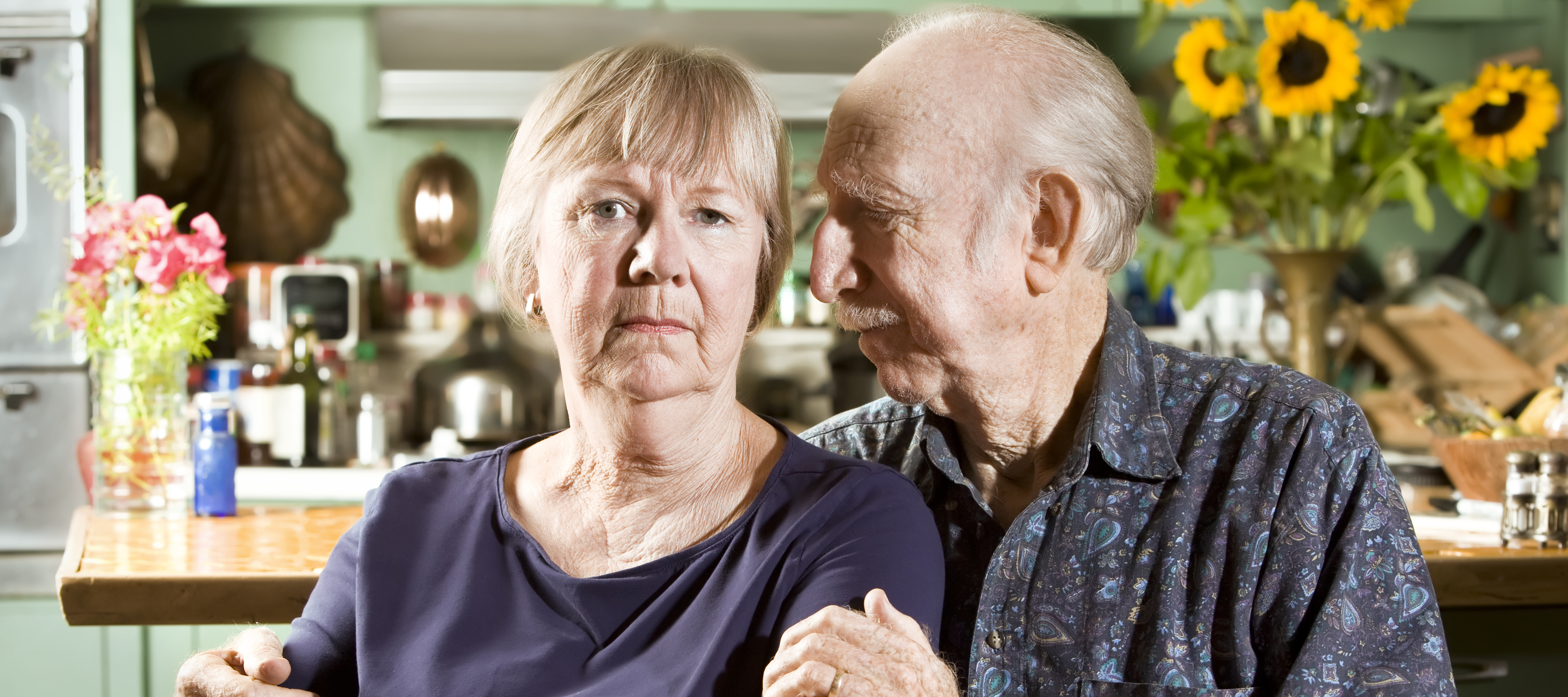 An older couple pose for a portrait at their kitchen table, lit by fading afternoon light.