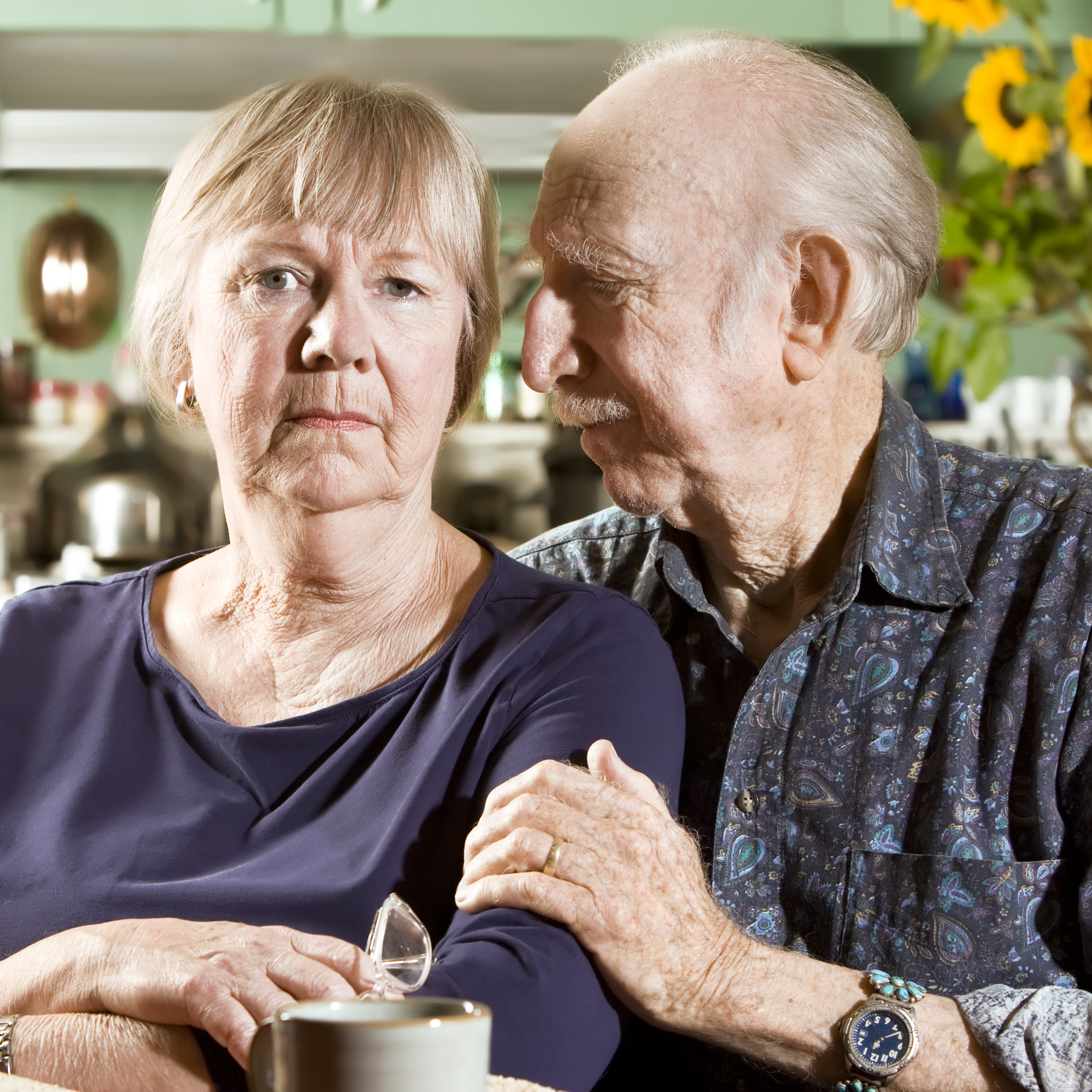An older couple pose for a portrait at their kitchen table, lit by fading afternoon light.