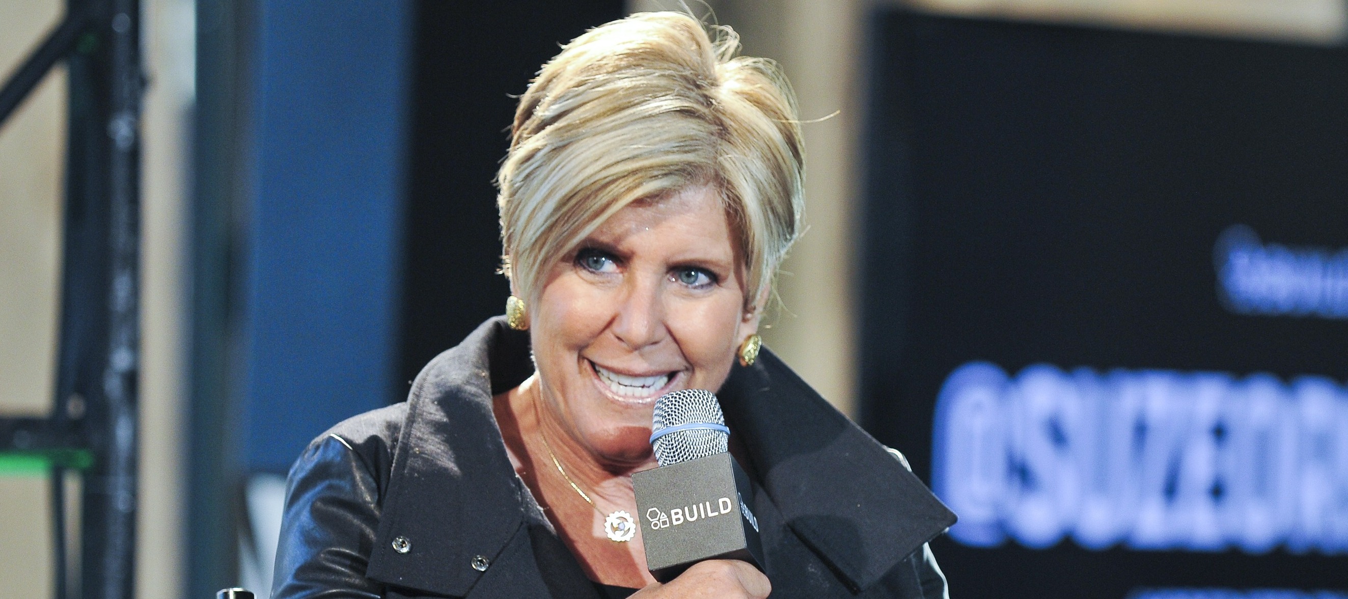 Suze Orman, in a black leather jacket, speaks into a microphone on stage.