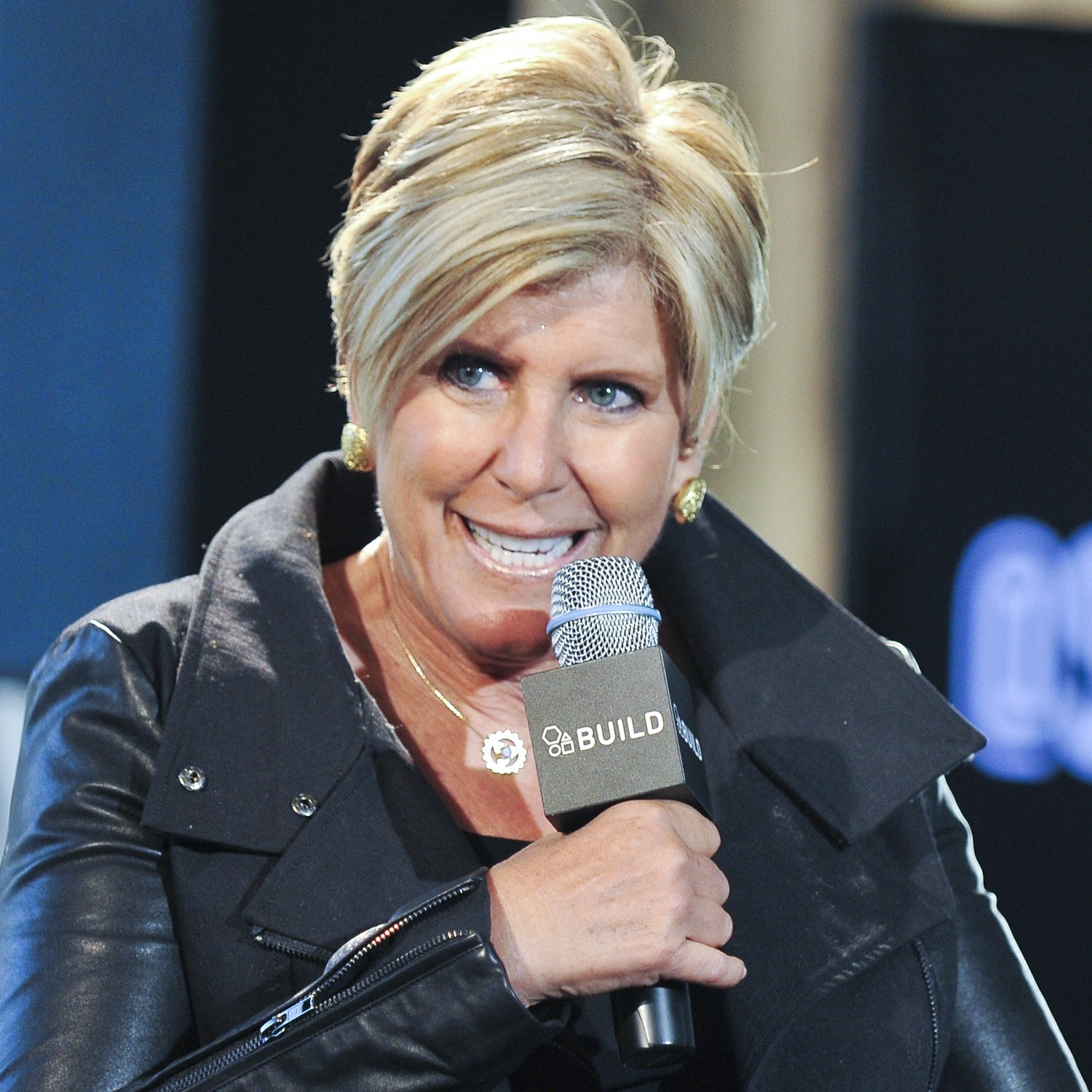 Suze Orman, in a black leather jacket, speaks into a microphone on stage.