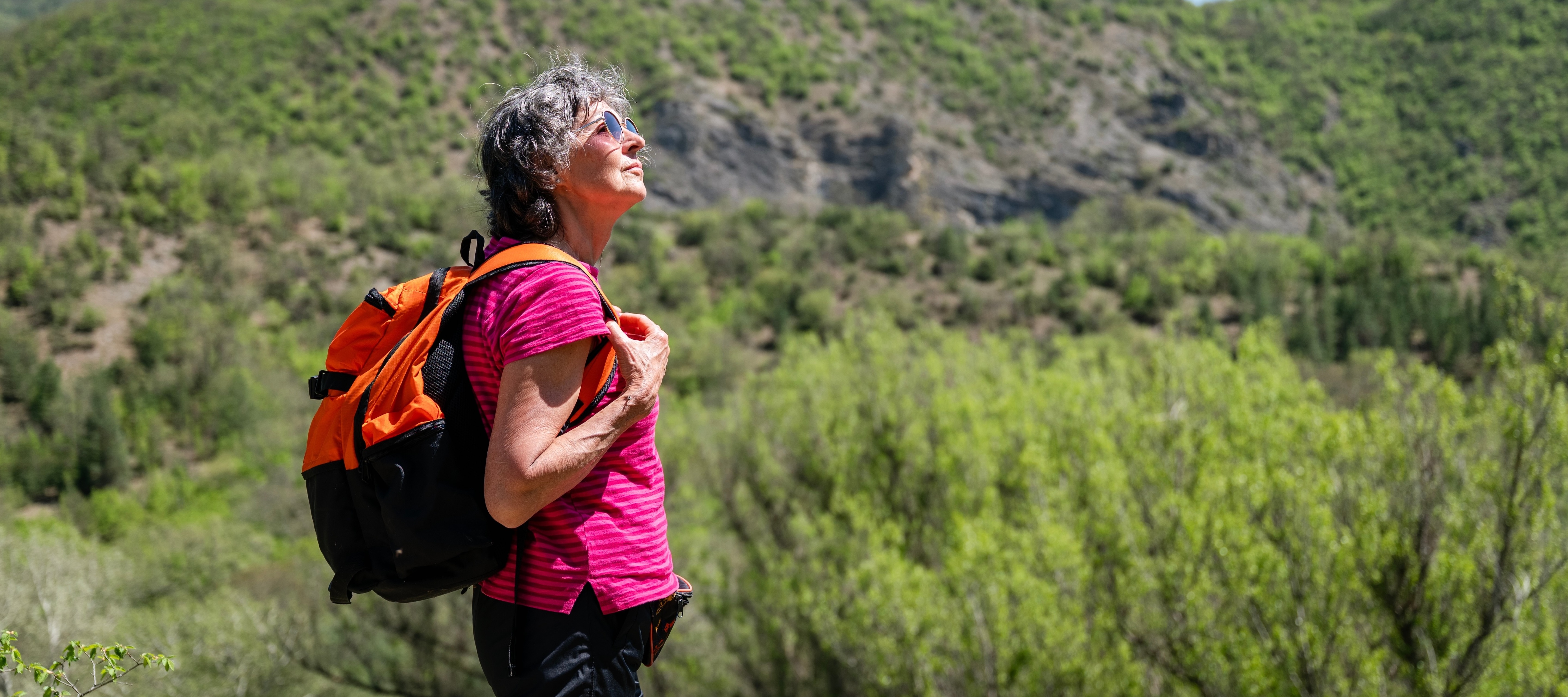 An older, gray-haired woman hiking in the summer, with rolling green hills in the background.