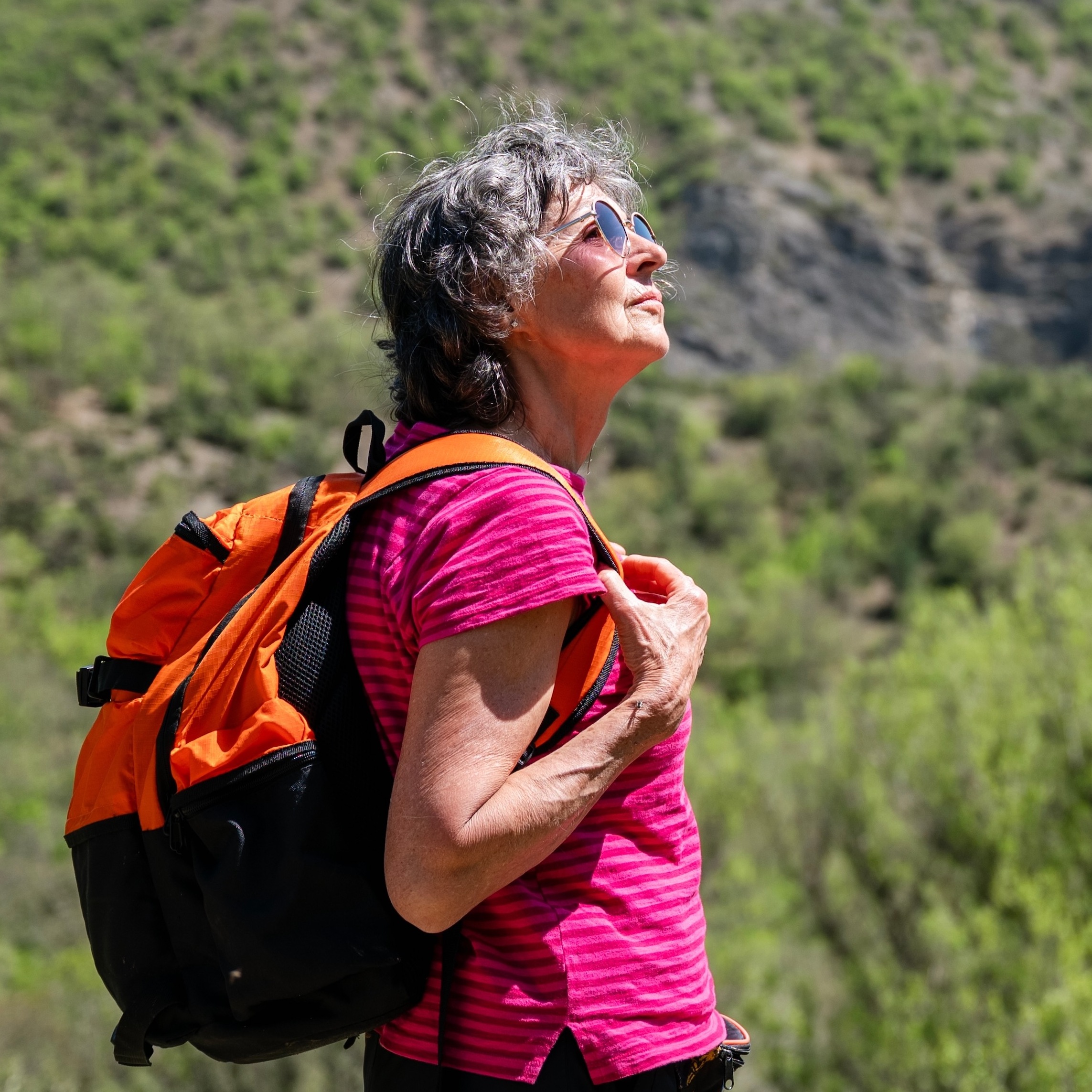 An older, gray-haired woman hiking in the summer, with rolling green hills in the background.