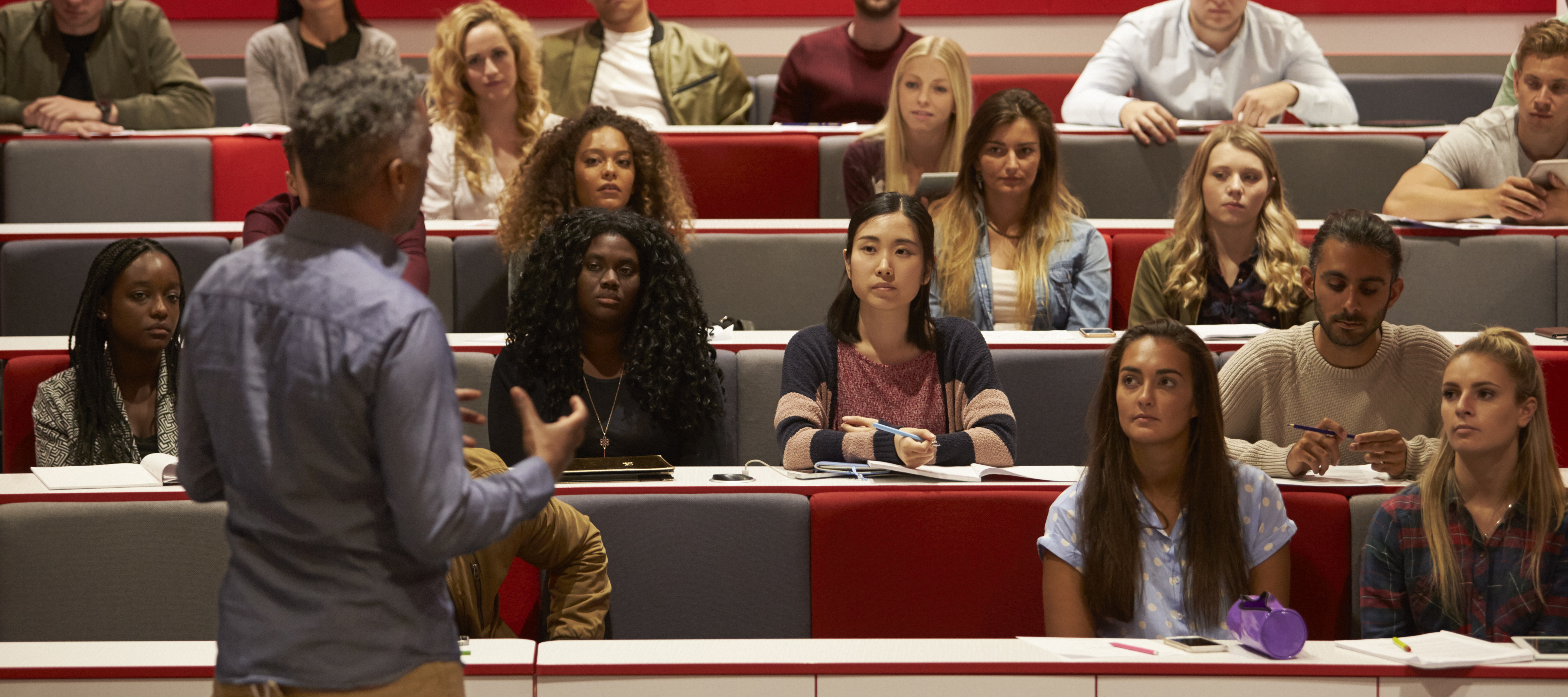 A group of students attentively listens to a professor in a lecture hall.