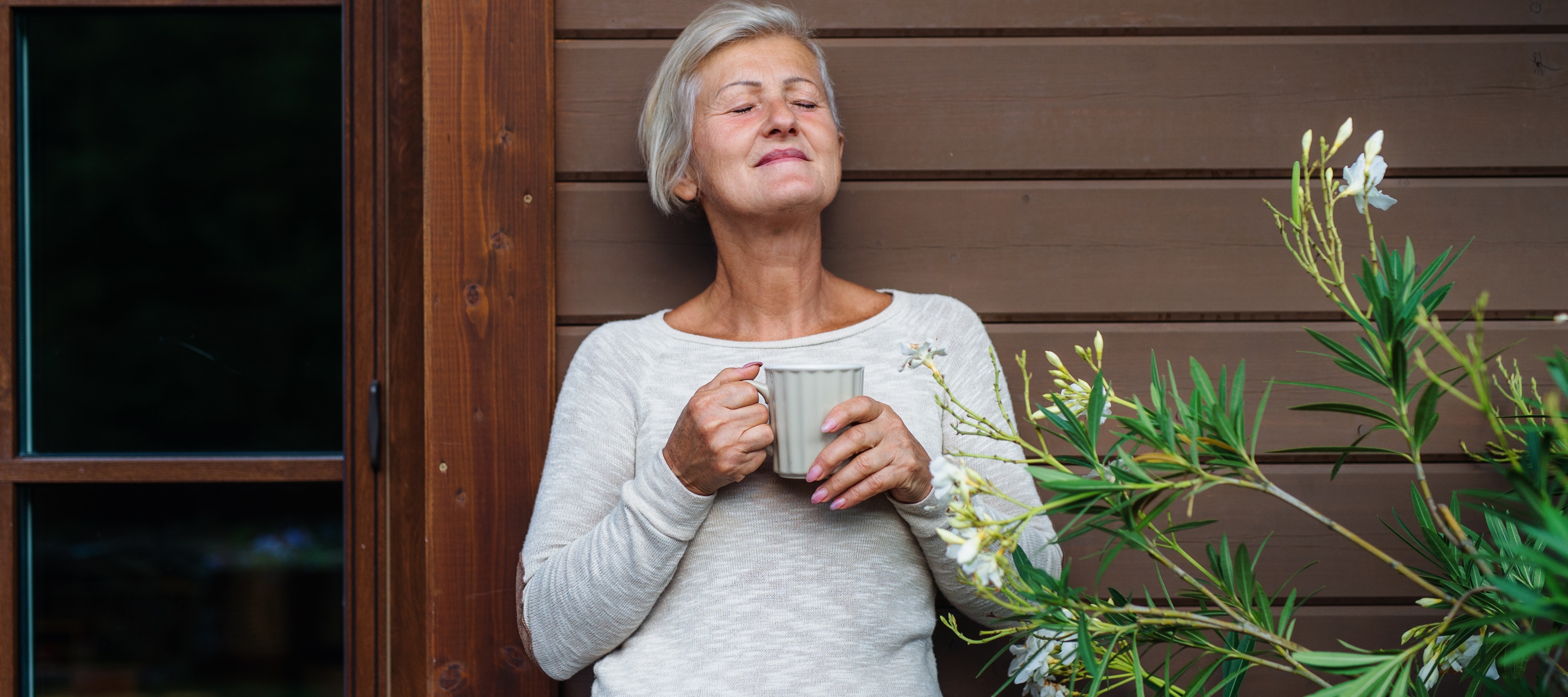 60-year-old woman drinking coffee and leaning against house on front porch