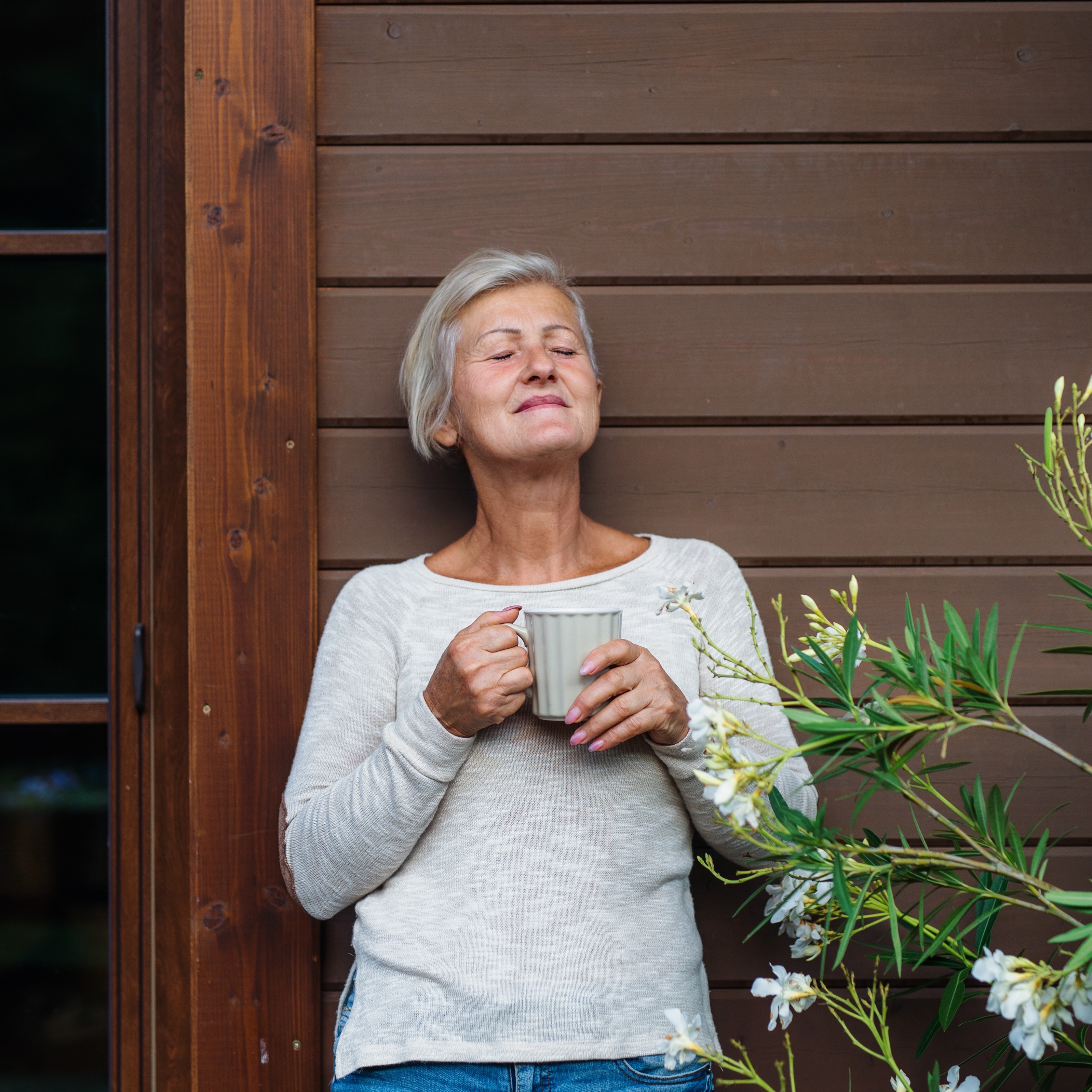 60-year-old woman drinking coffee and leaning against house on front porch