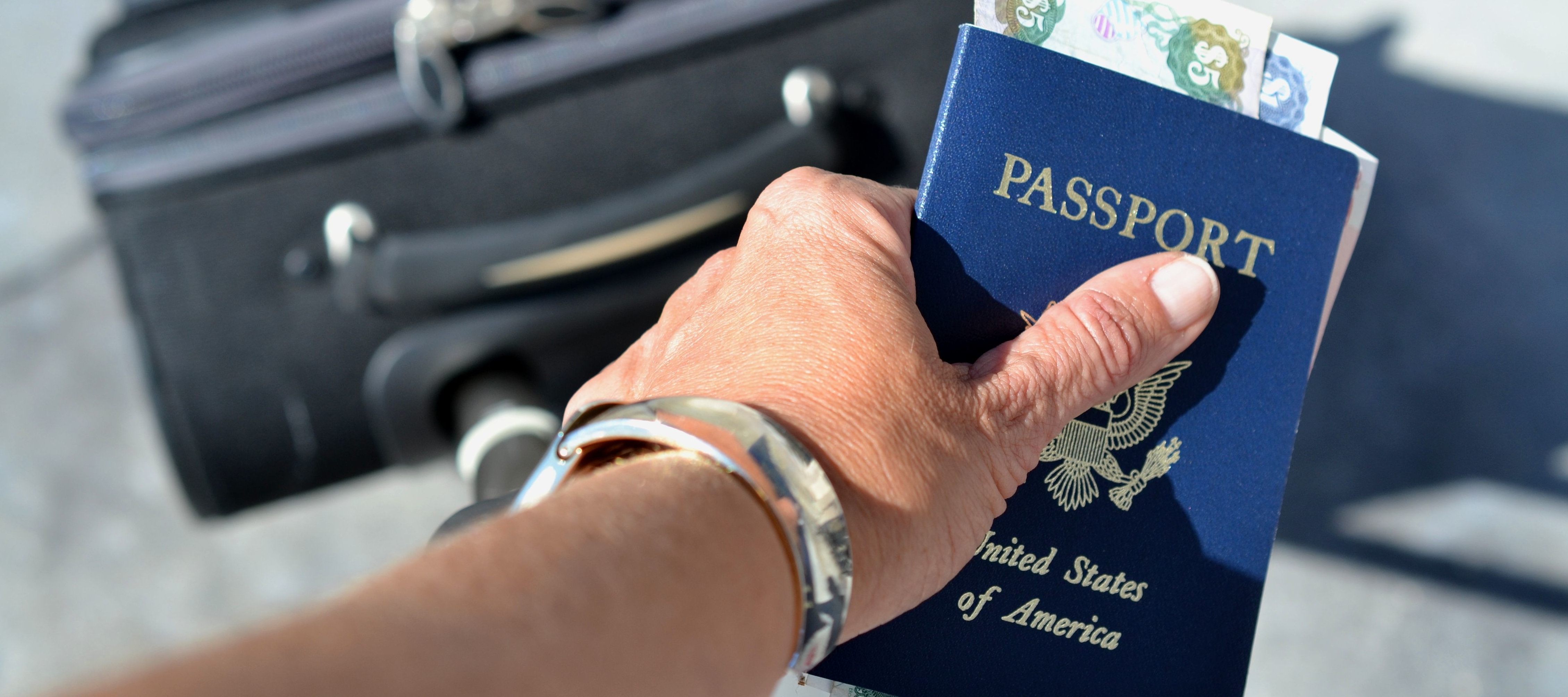 Woman's hand holding U.S. passport, suitcase handle and foreign currency