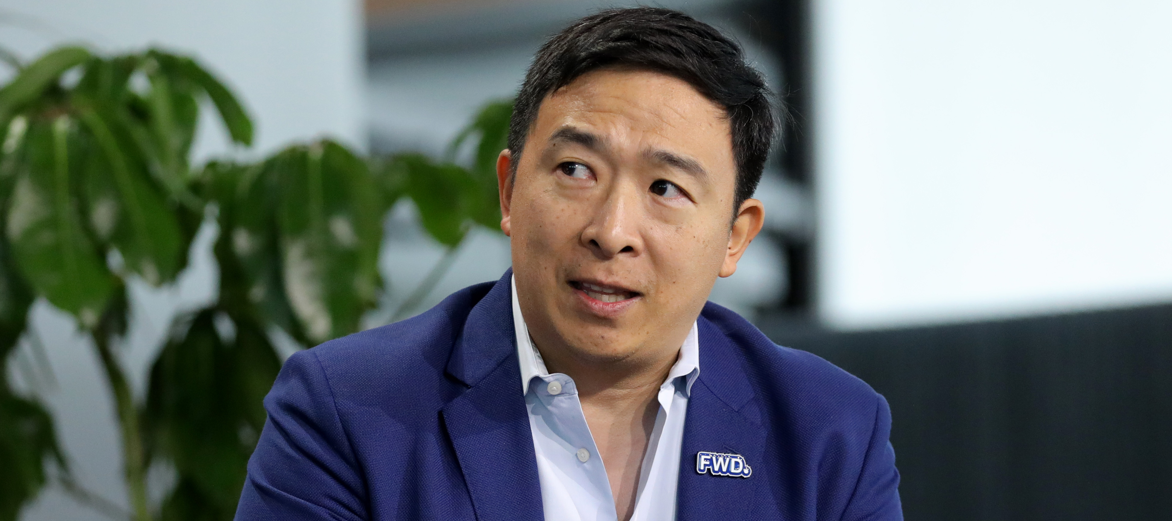 Andrew Yang raises one hand, looking concerned, while speaking on stage in a black suit jacket.