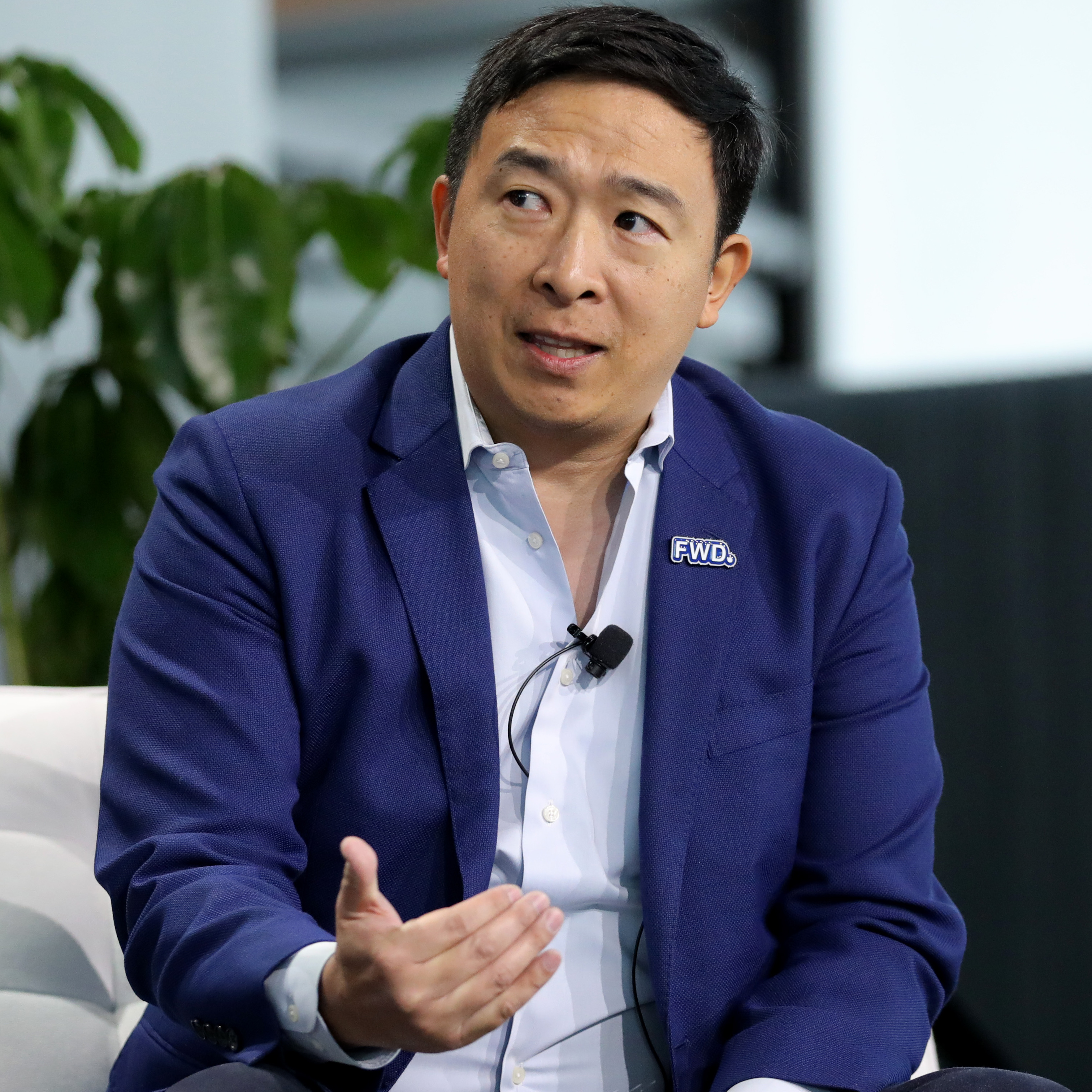 Andrew Yang raises one hand, looking concerned, while speaking on stage in a black suit jacket.
