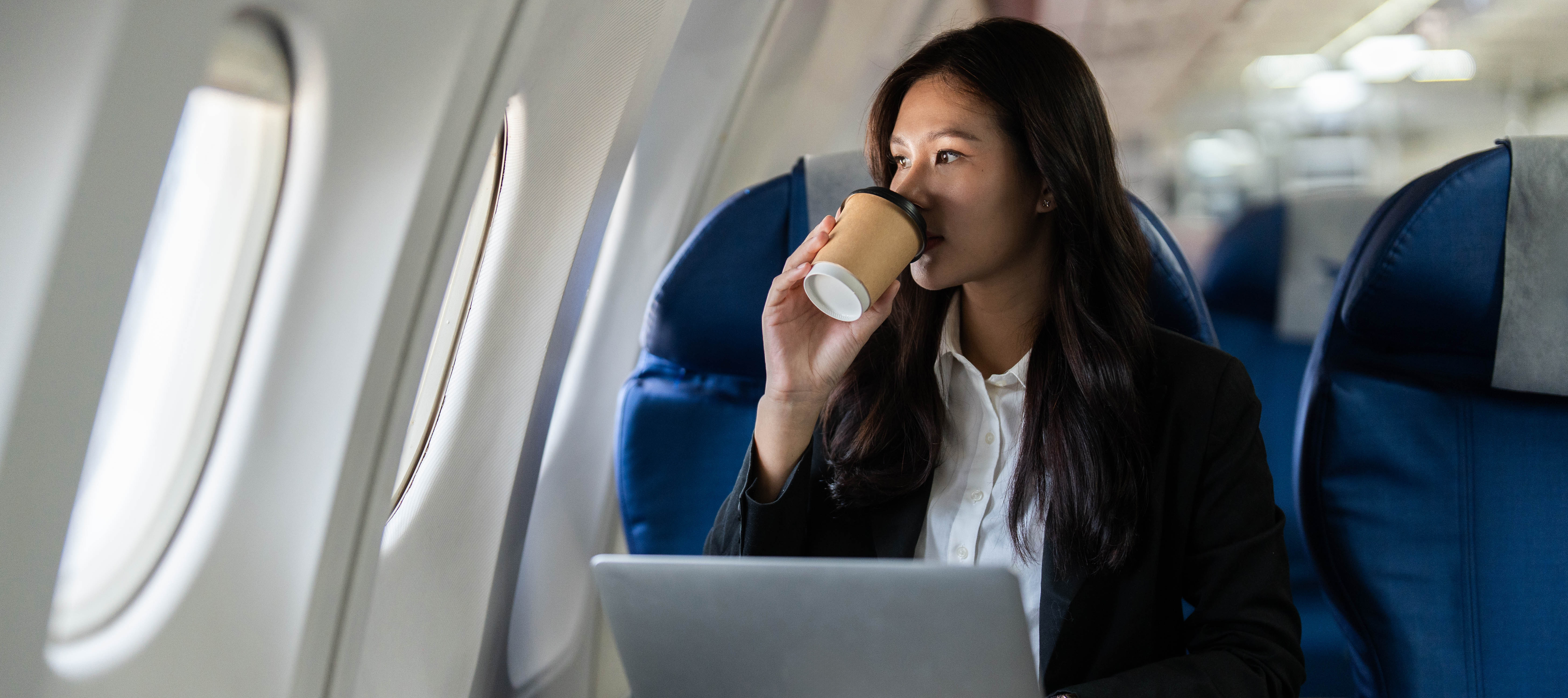 A young adult woman with long dark hair sits in a window seat on an airplane.