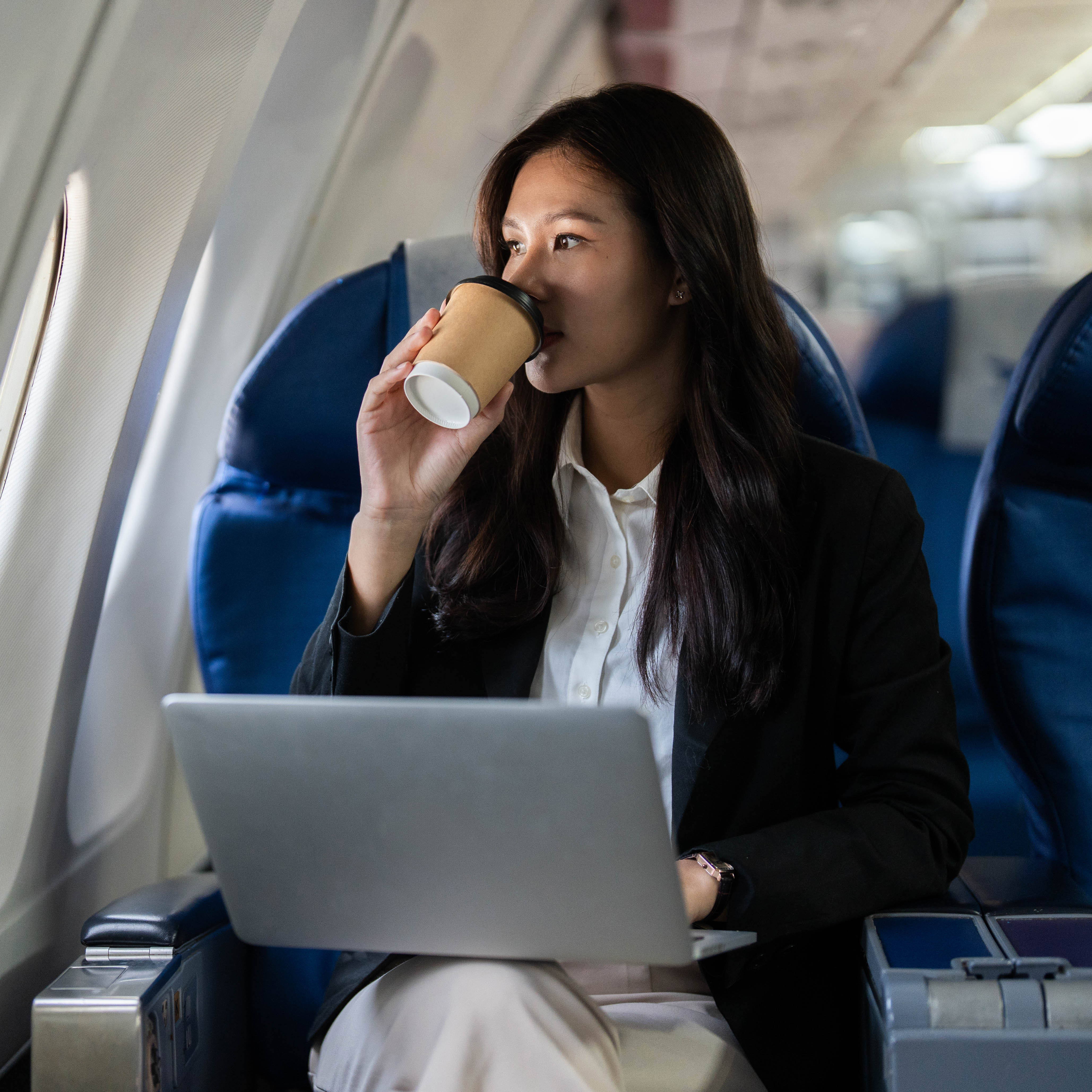 A young adult woman with long dark hair sits in a window seat on an airplane.