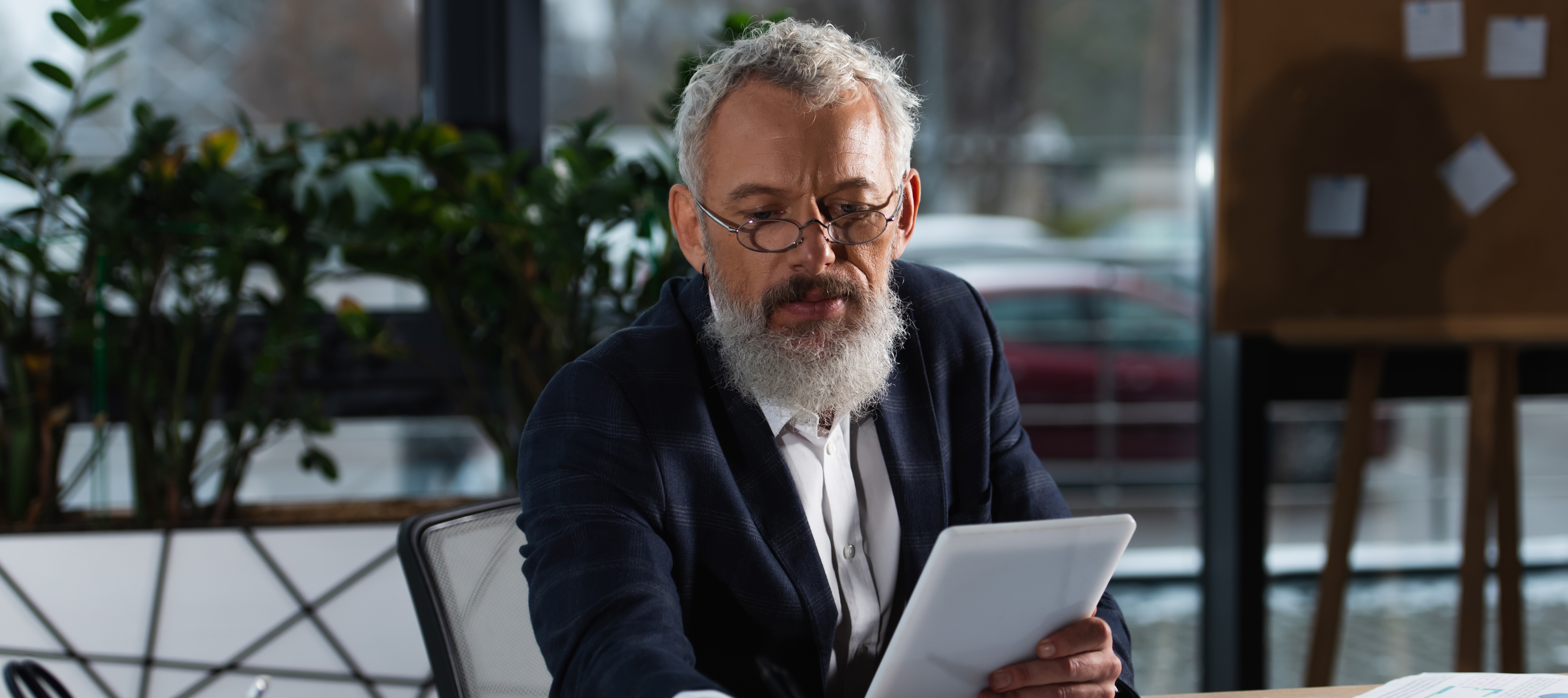 A man with white hair and a beard wearing glasses and a blazer sits at a desk holding a tablet in one hand and a pen in the other while reviewing documents.