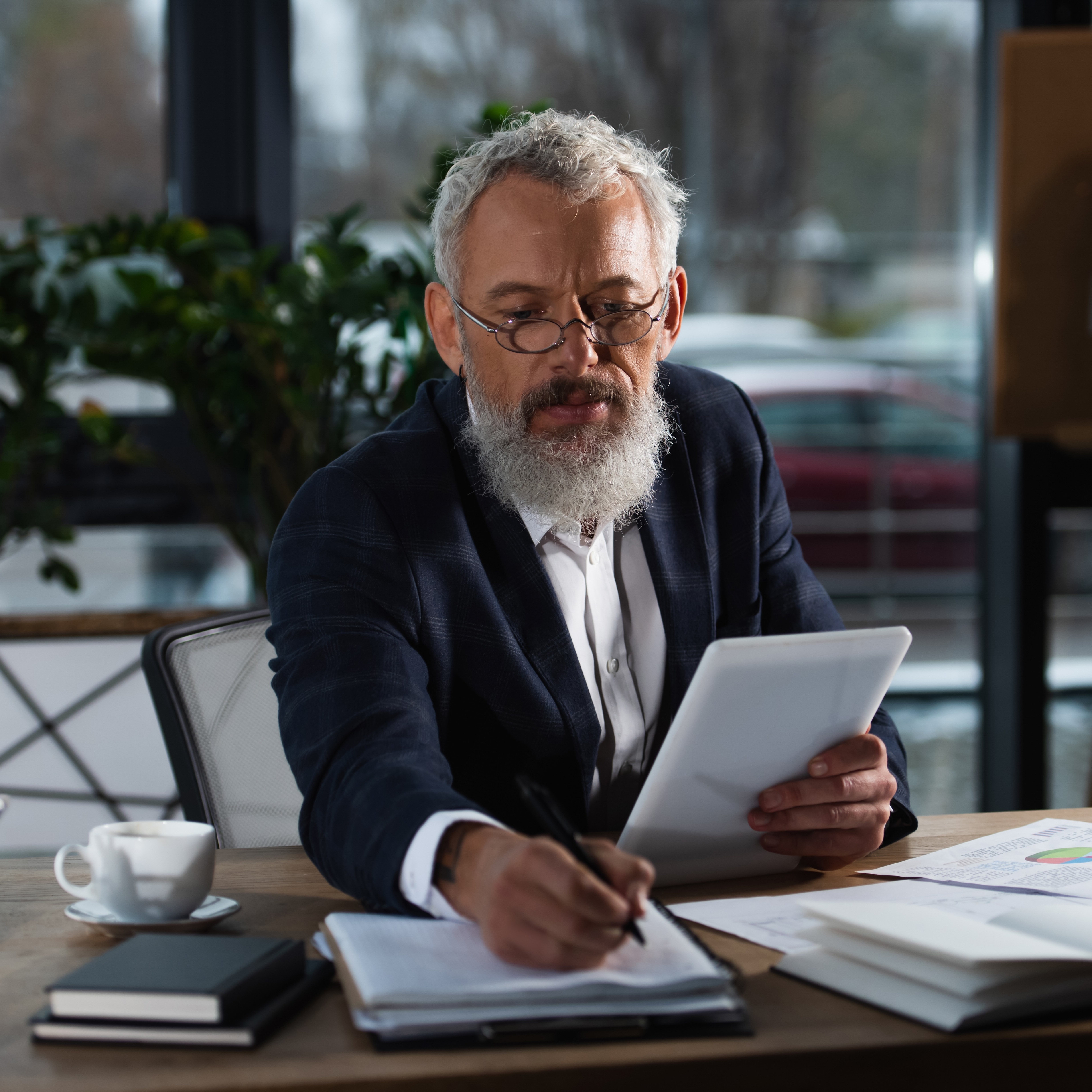 A man with white hair and a beard wearing glasses and a blazer sits at a desk holding a tablet in one hand and a pen in the other while reviewing documents.