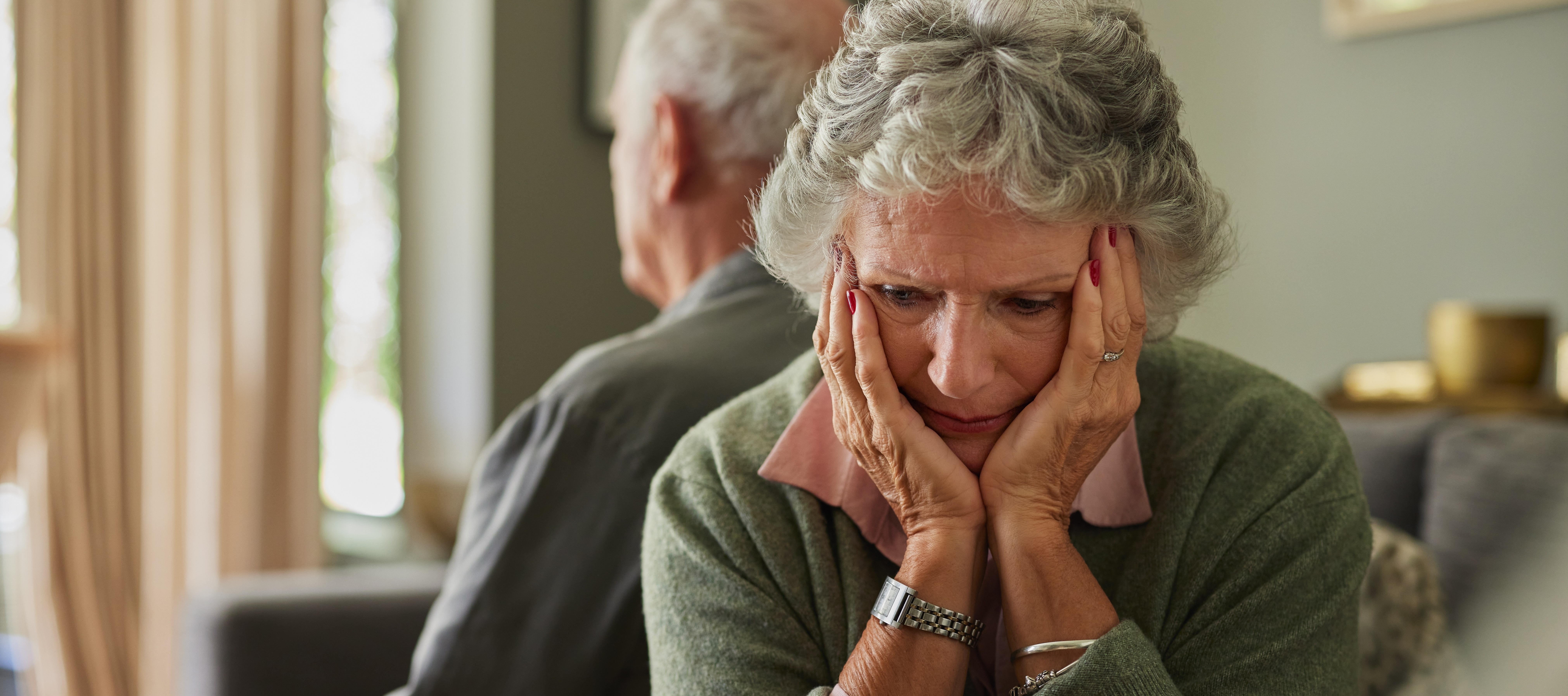A focused waist-up shot shows a senior woman in a green sweater sitting indoors, her hands placed on her face in what appears to be a moment of sadness.