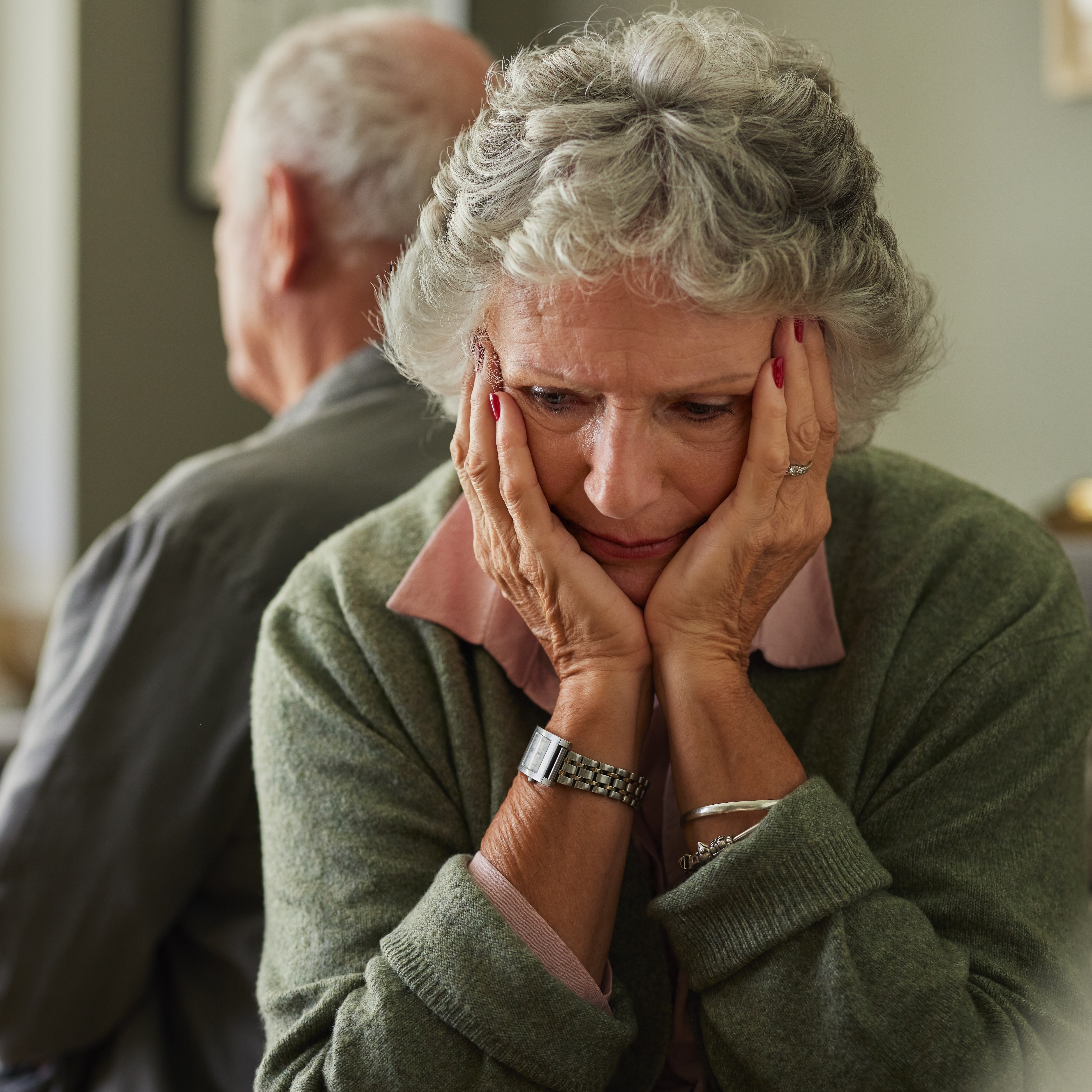 A focused waist-up shot shows a senior woman in a green sweater sitting indoors, her hands placed on her face in what appears to be a moment of sadness.