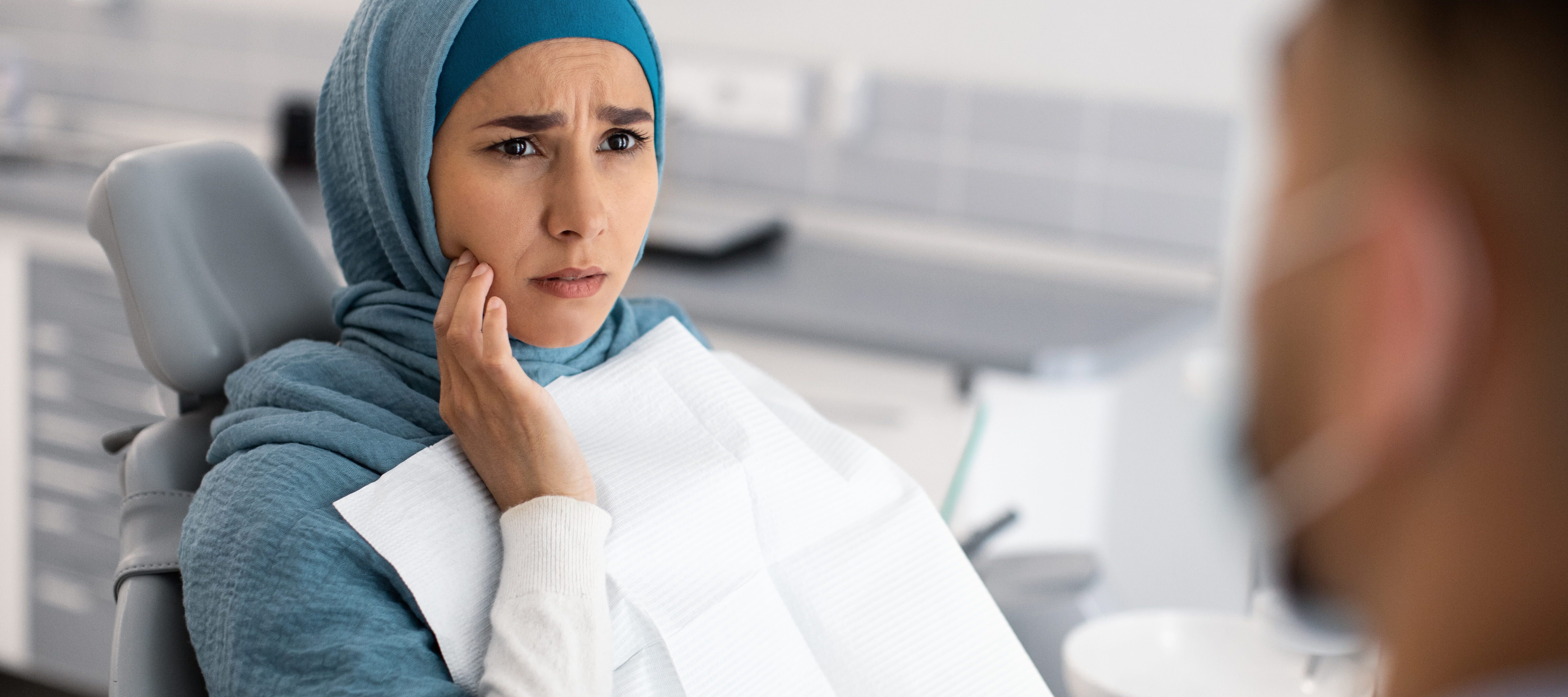 Young woman wearing blue headscarf looking concerned at dentist