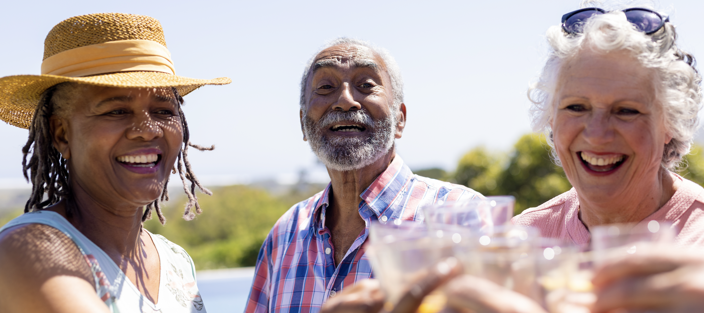 Three senior adults smile and raise glasses in a toast on a bright, sunny day.