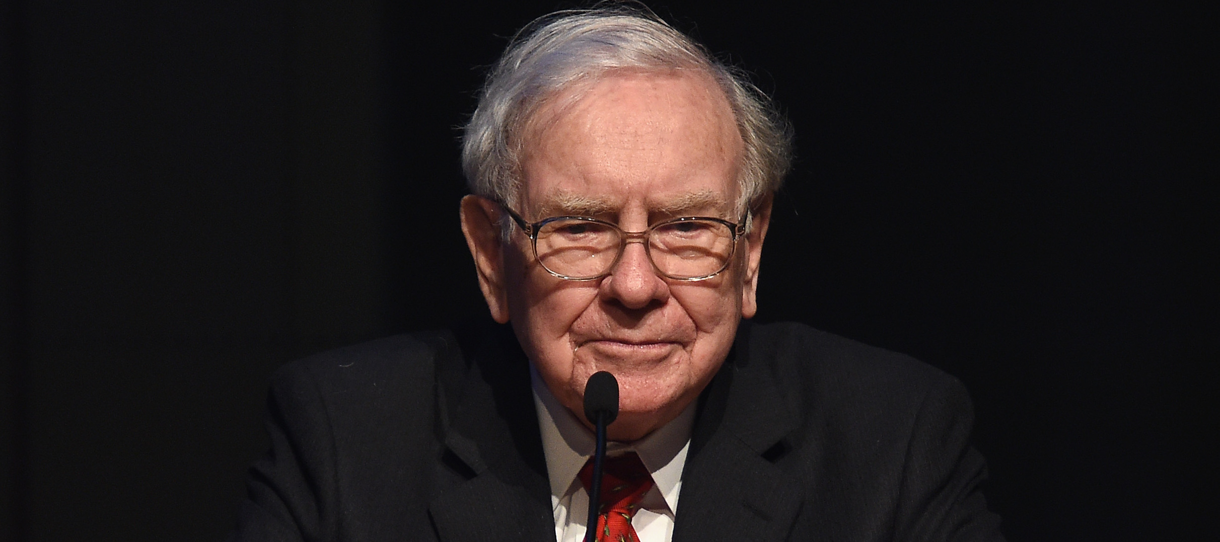 Warren Buffett stands at a podium, looking grim with a stark black background behind him.