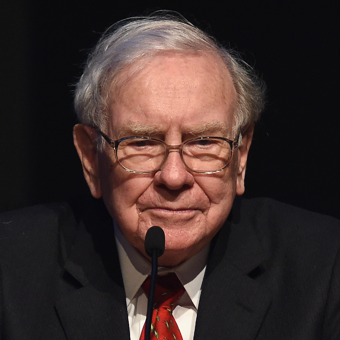 Warren Buffett stands at a podium, looking grim with a stark black background behind him.