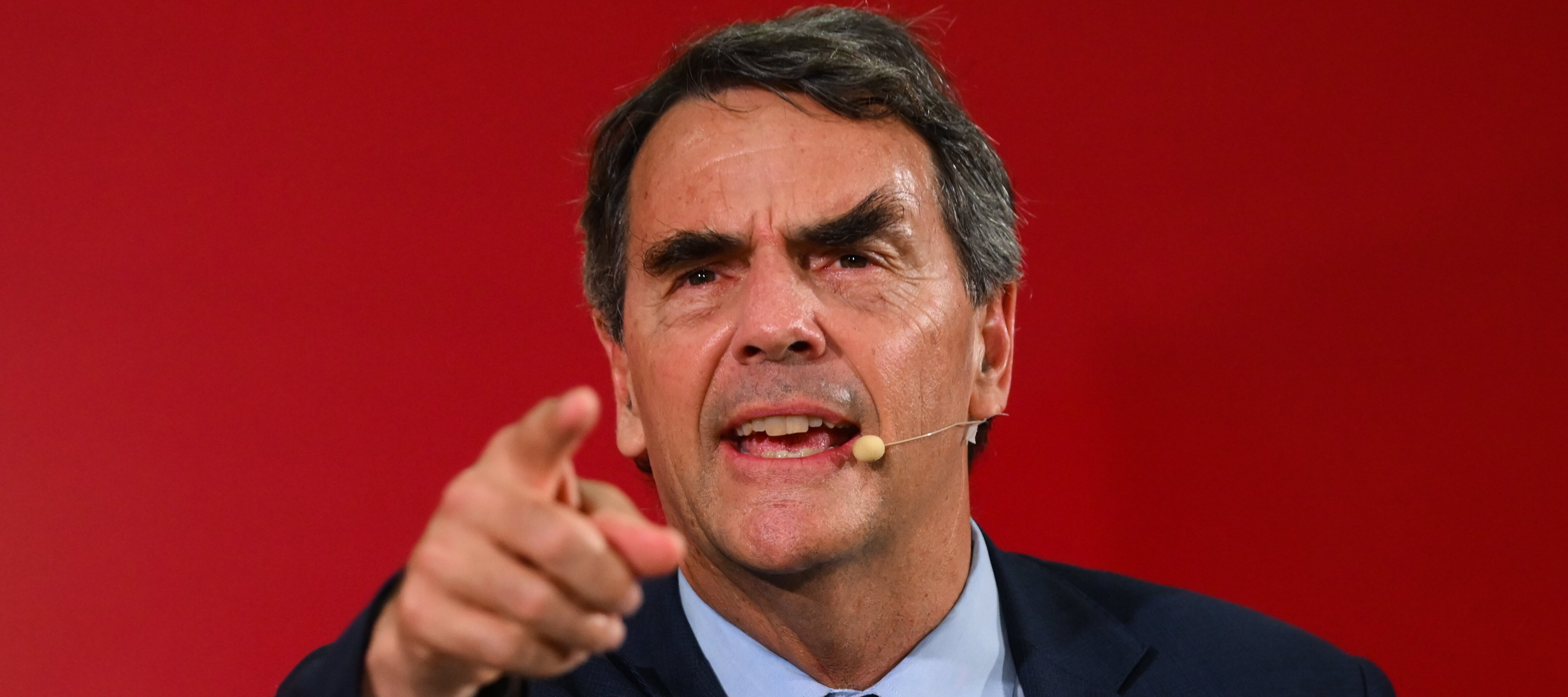 Tim Draper, wearing his iconic purple tie with dollar coin prints, points directly at the viewer while answering a question on stage.