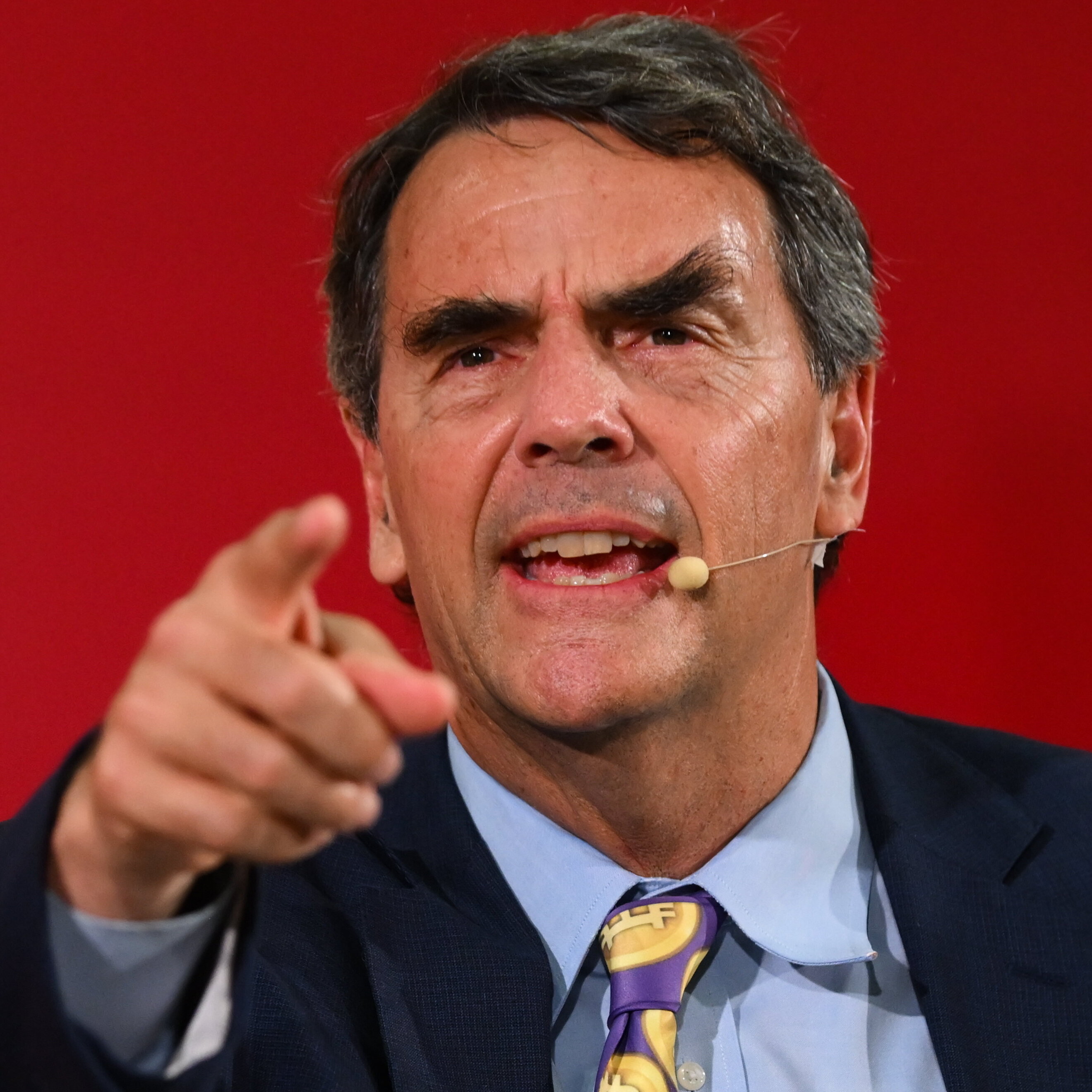 Tim Draper, wearing his iconic purple tie with dollar coin prints, points directly at the viewer while answering a question on stage.