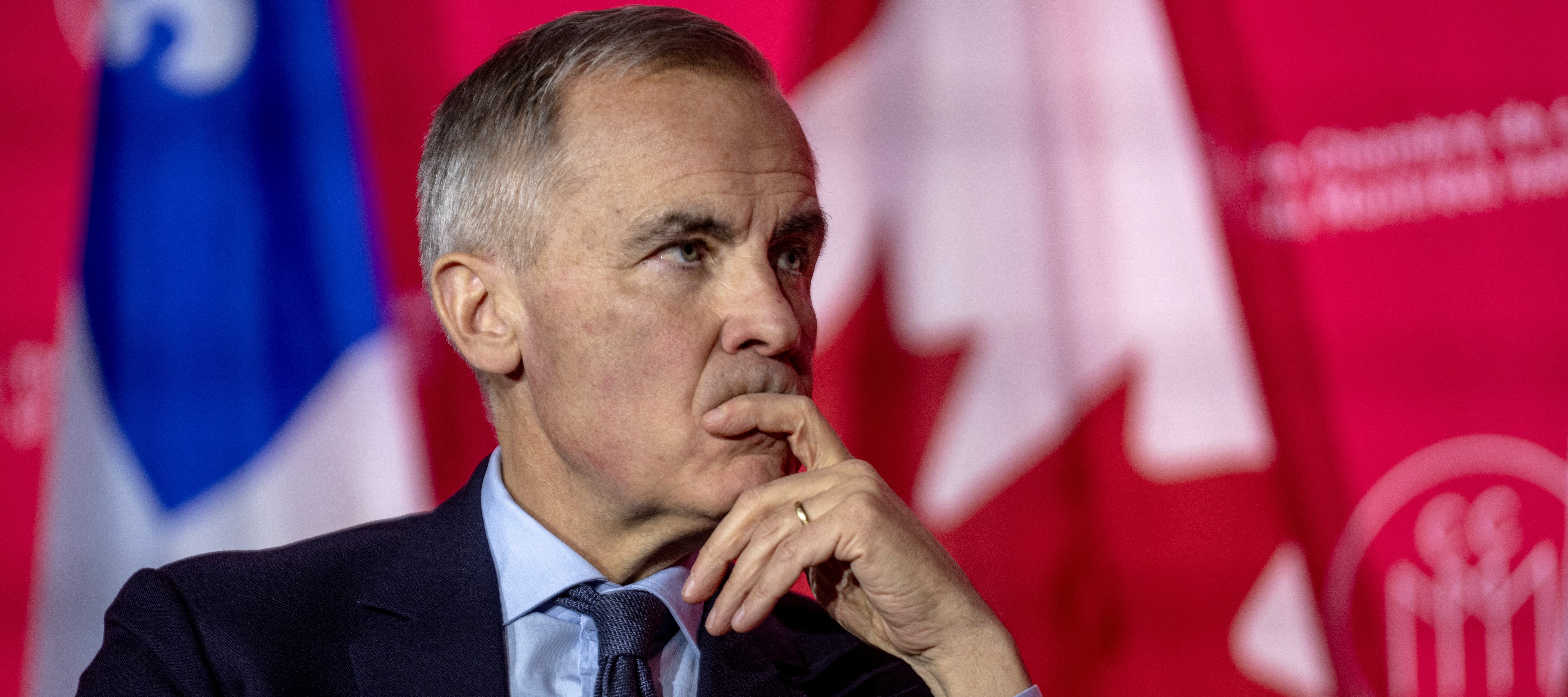Prime Minister Mark Carney sits on stage with one hand covering his mouth, staring into the distance.