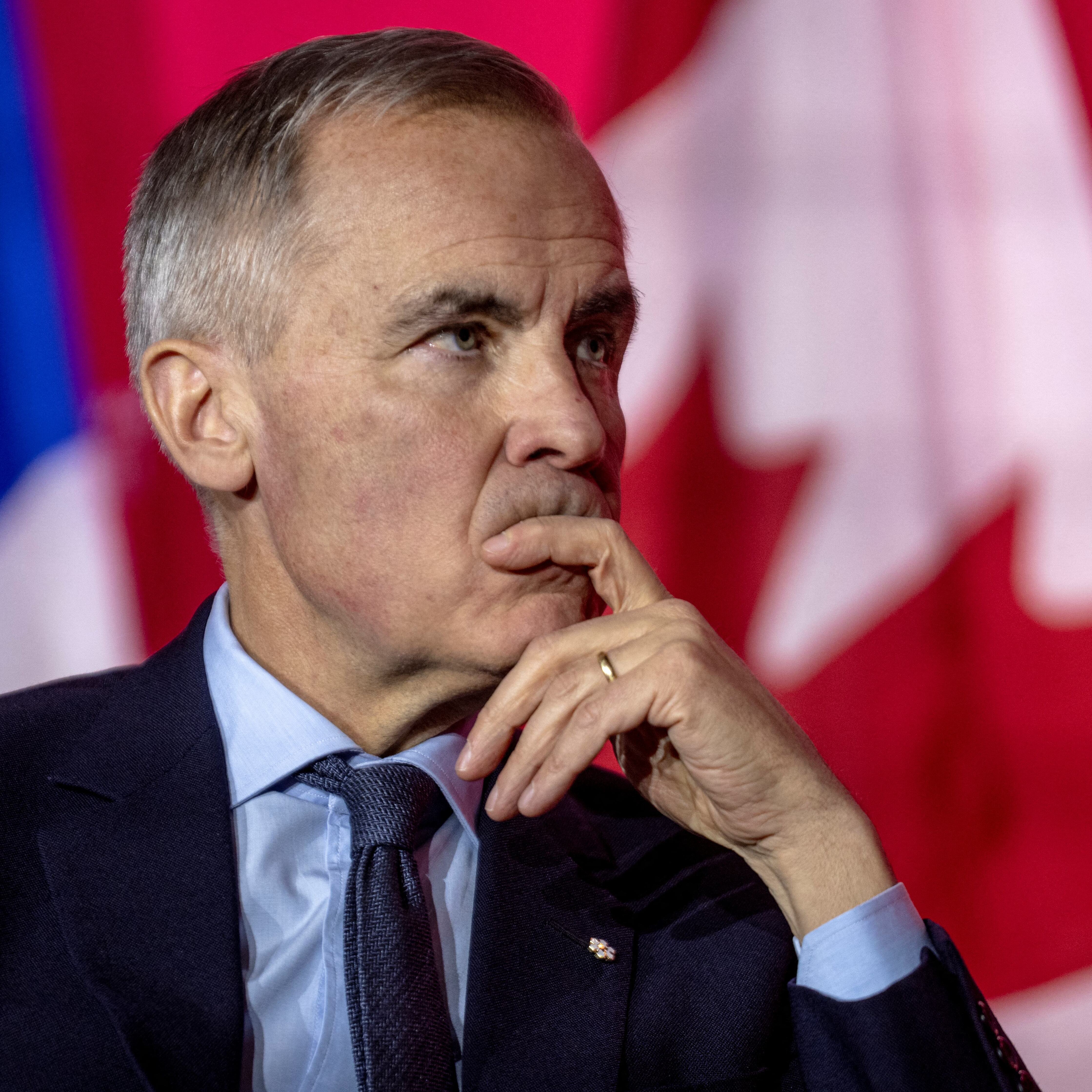 Prime Minister Mark Carney sits on stage with one hand covering his mouth, staring into the distance.