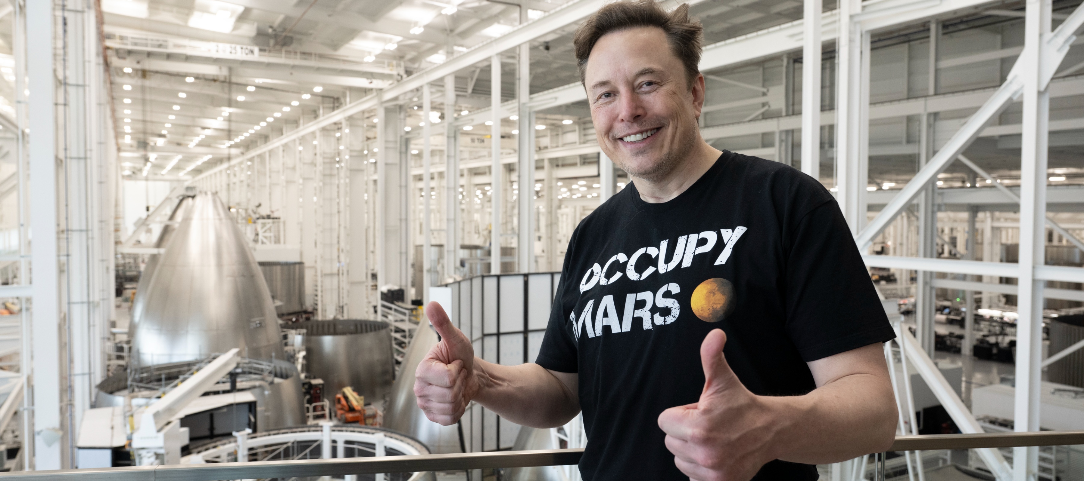 Elon Musk gives two thumbs up as he stands inside of a SpaceX facility