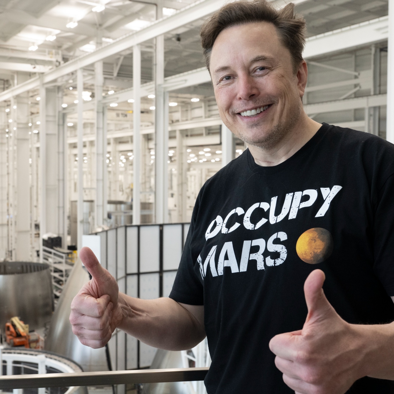 Elon Musk gives two thumbs up as he stands inside of a SpaceX facility