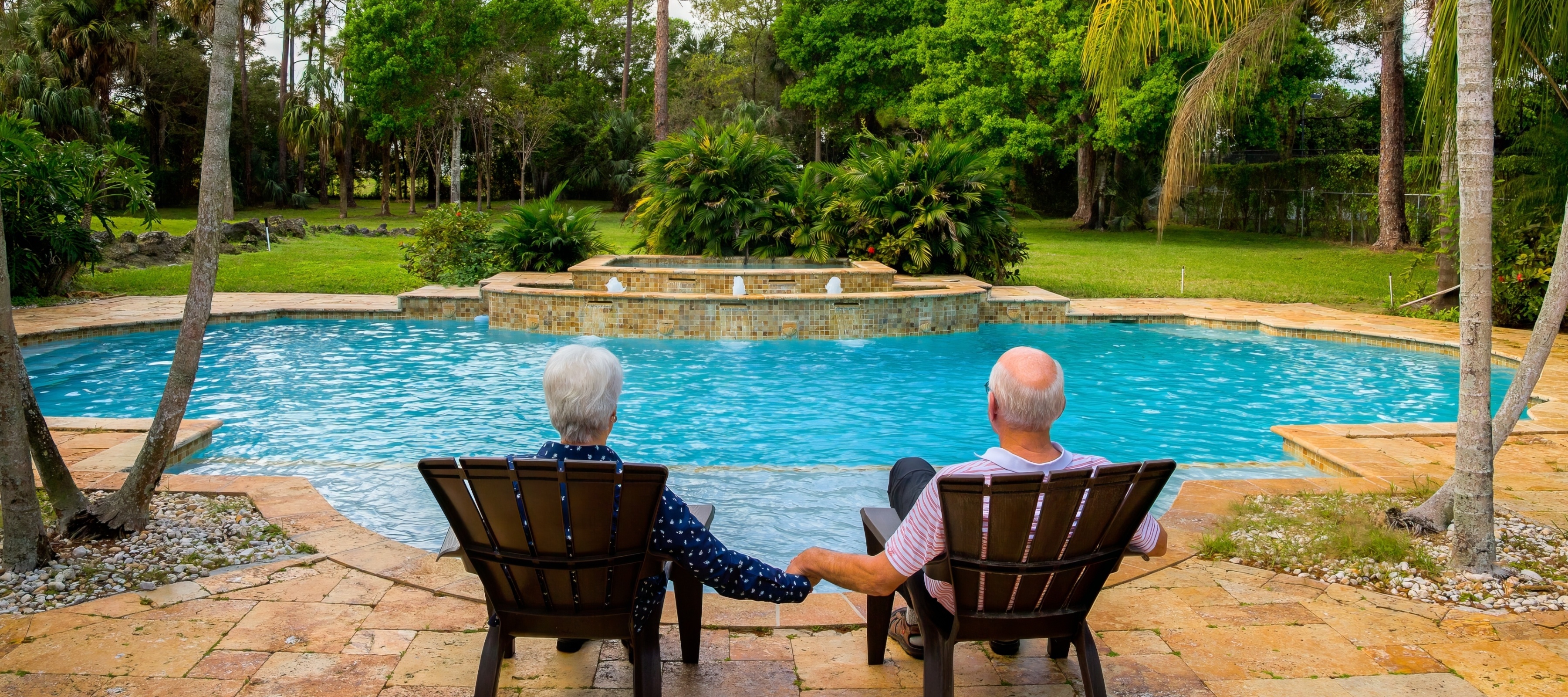 An old retired couple holding hands in beach chairs sitting in front of a pool surrounded by palm trees