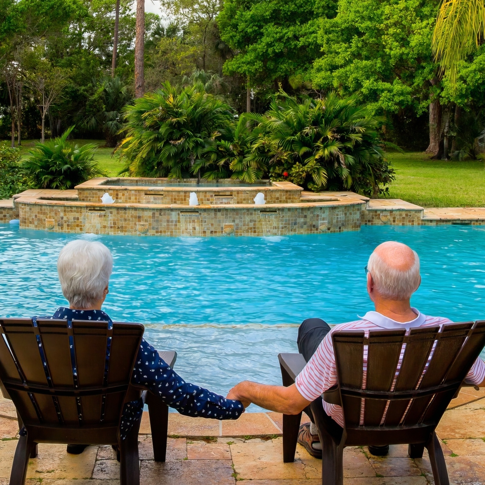 An old retired couple holding hands in beach chairs sitting in front of a pool surrounded by palm trees