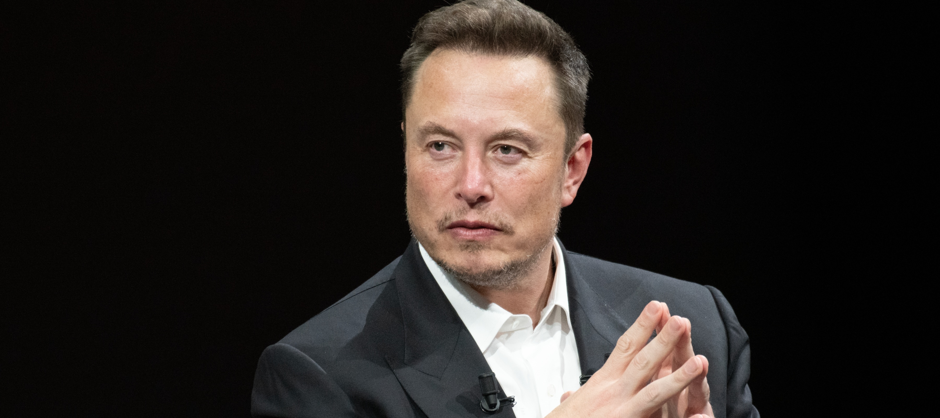 Elon Musk sits in a chair with his hands folded