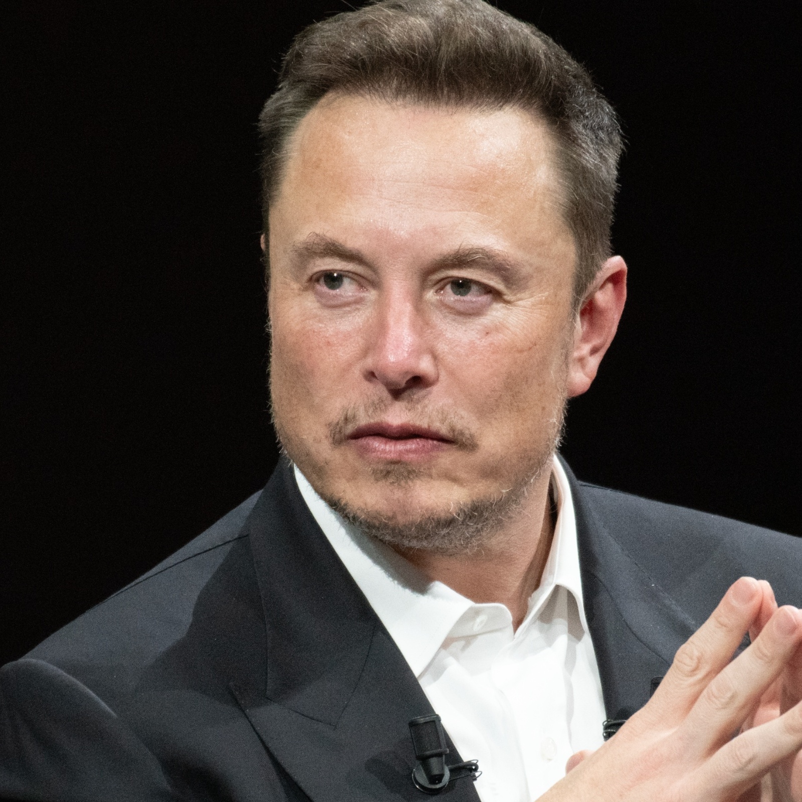 Elon Musk sits in a chair with his hands folded