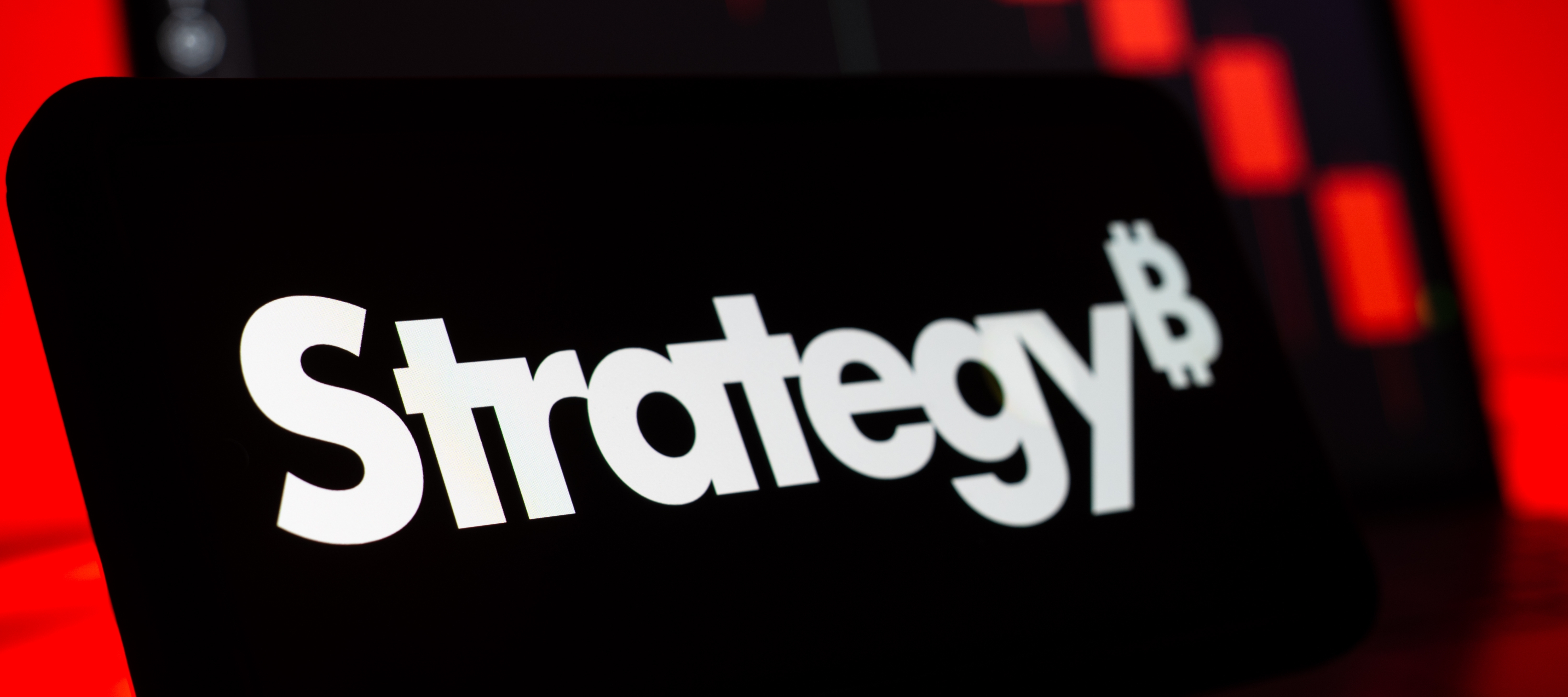 A close up of the Strategy logo on a smartphone with candlestick investing patterns in the background.