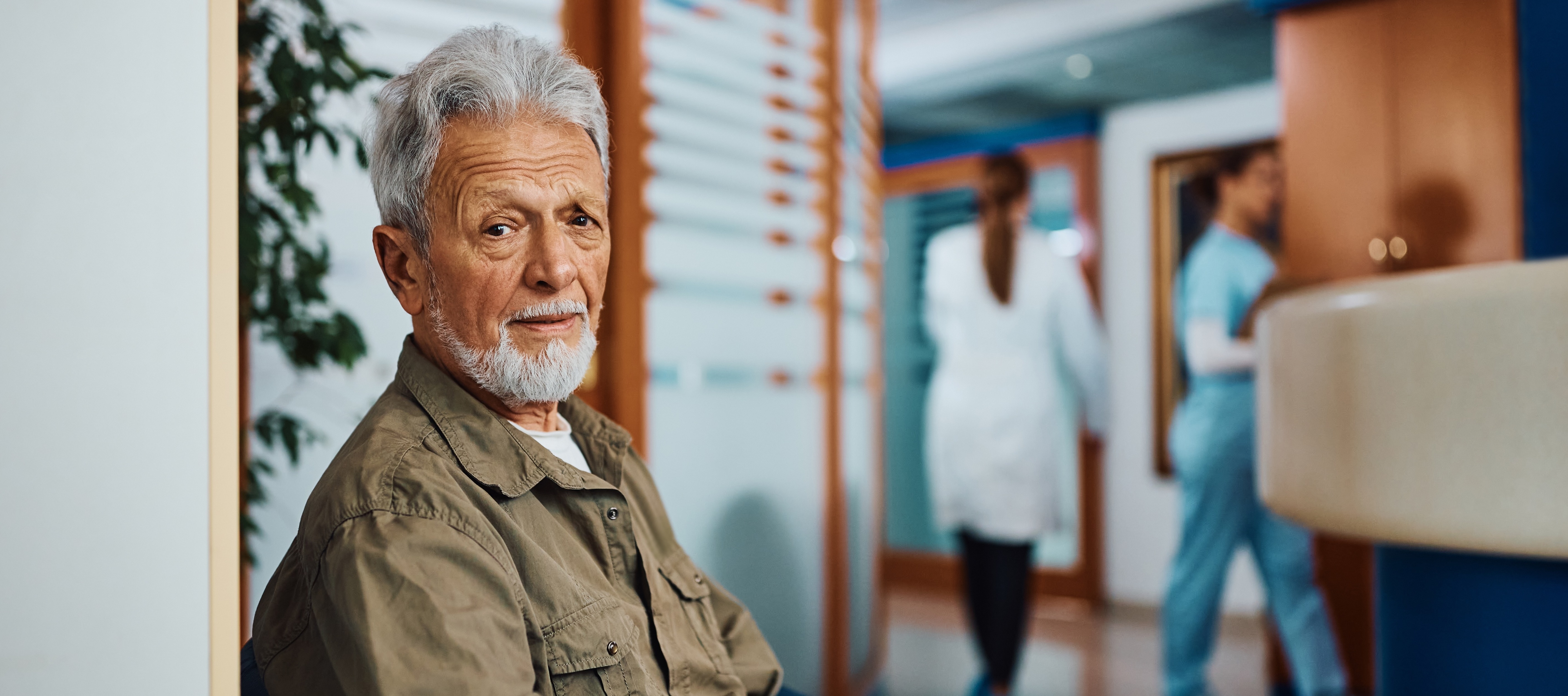 In a bright waiting room, a mature man with gray hair and a beard sits patiently, looking directly at the camera while holding paperwork.