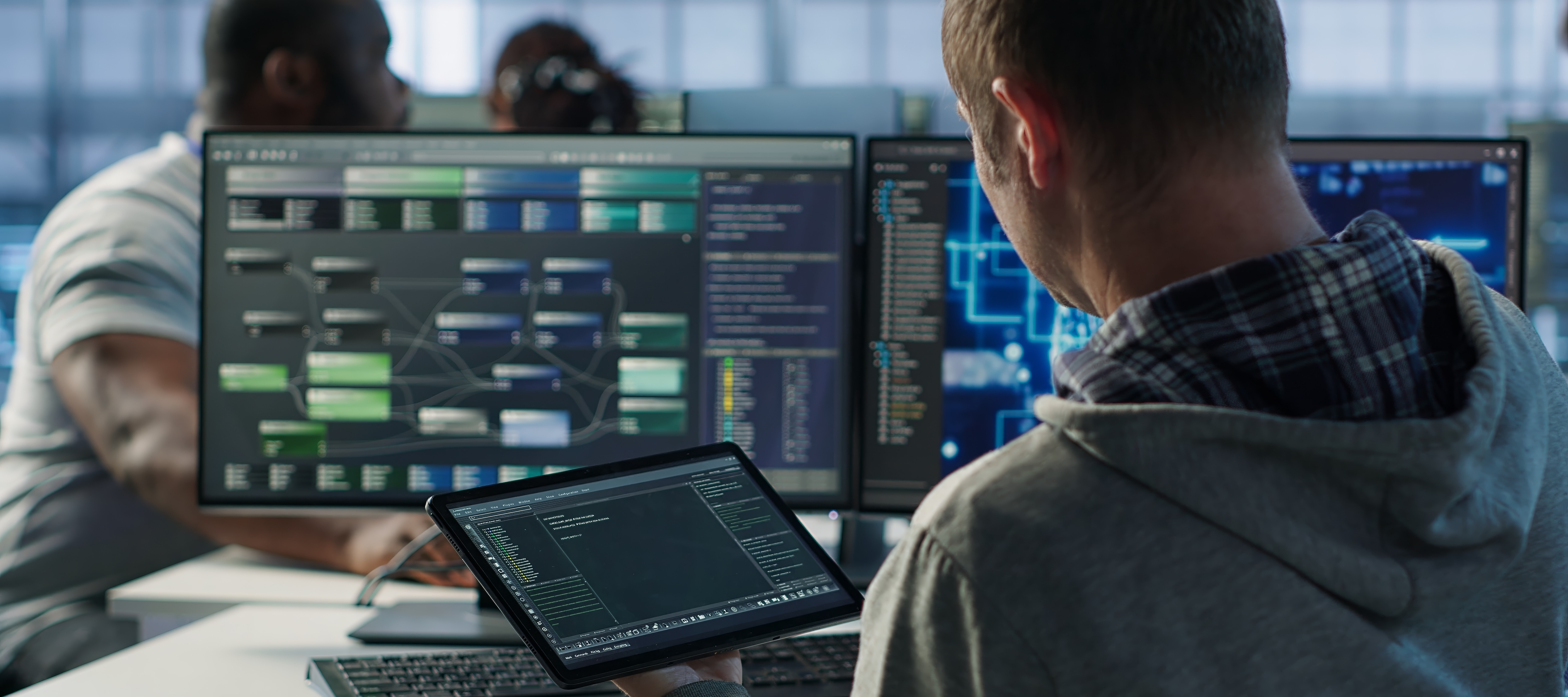 The back side view of a person working on a computer and tablet with multiple screens and programs running at once.