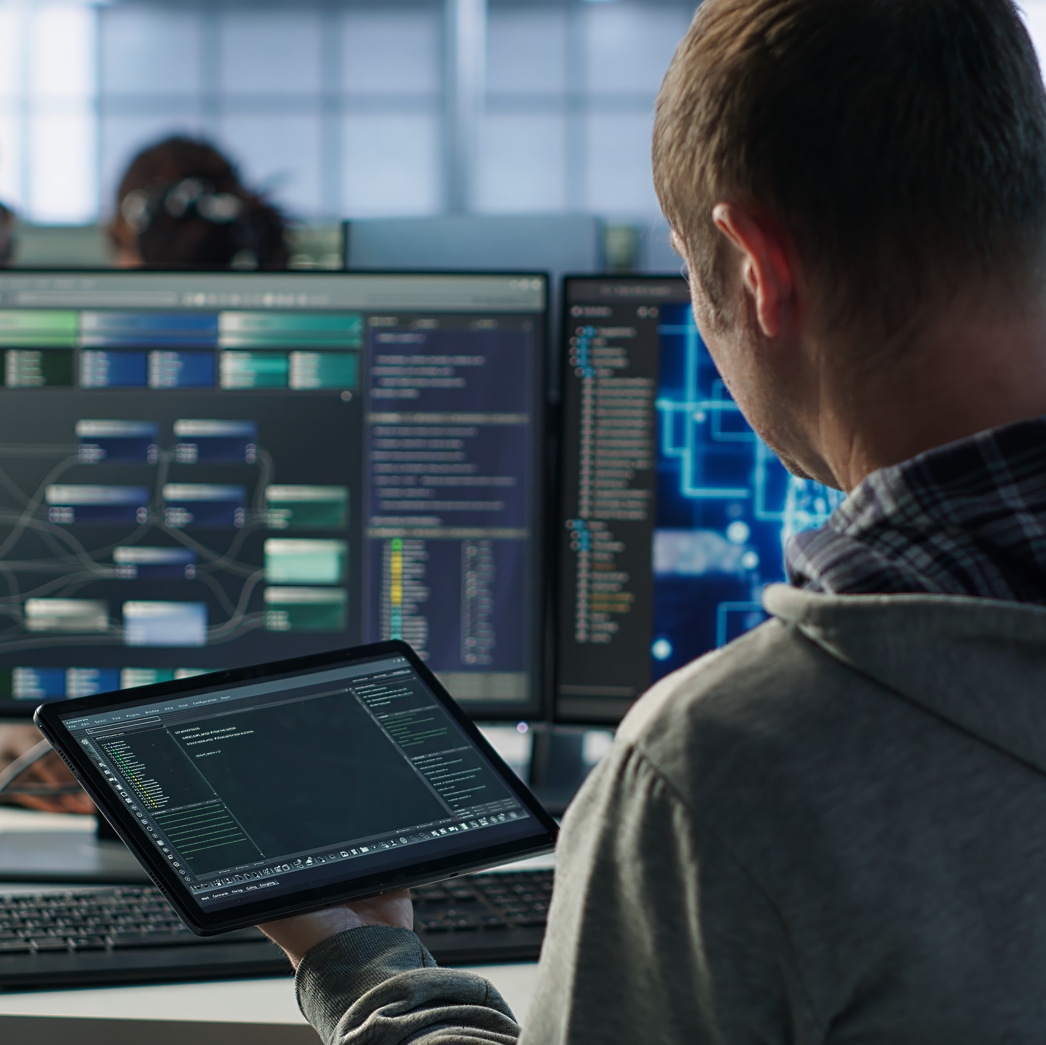 The back side view of a person working on a computer and tablet with multiple screens and programs running at once.