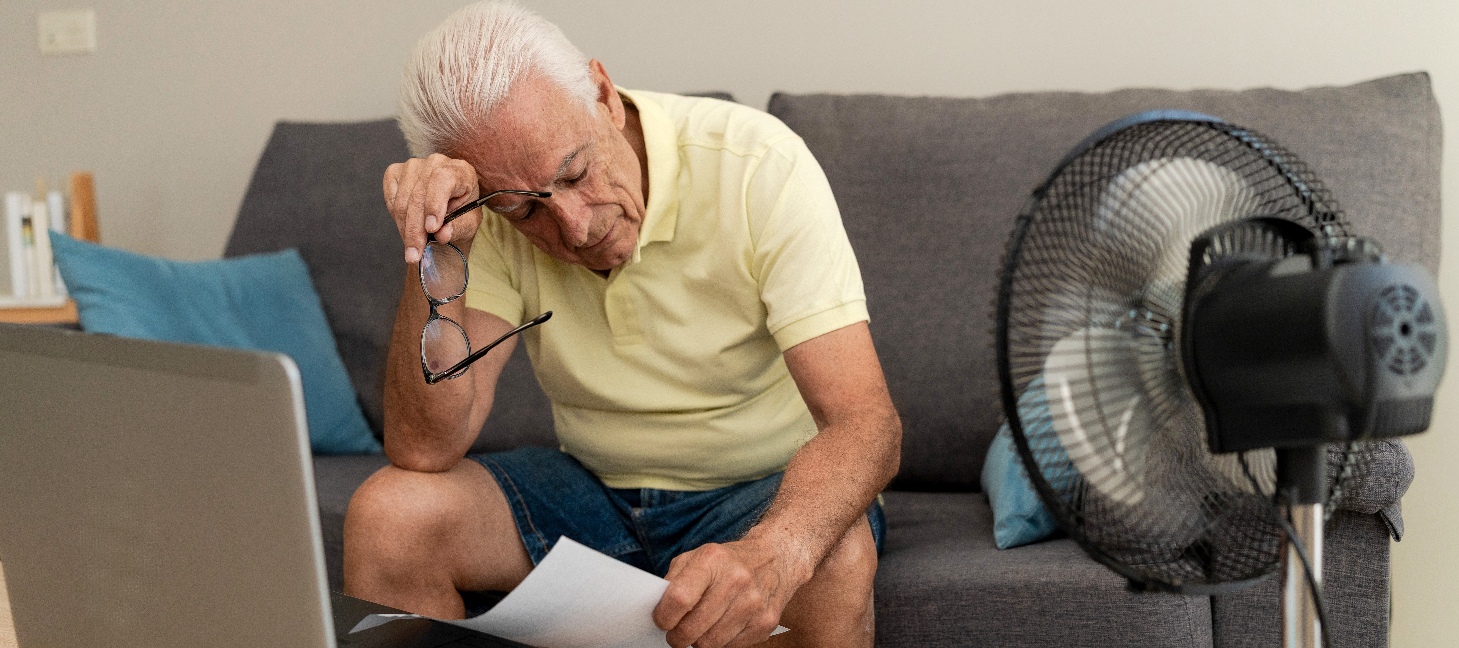 An older man sits on a couch with a fan close by blowing on him. A laptop sits on the coffee table in front of him and he wipes his brow.