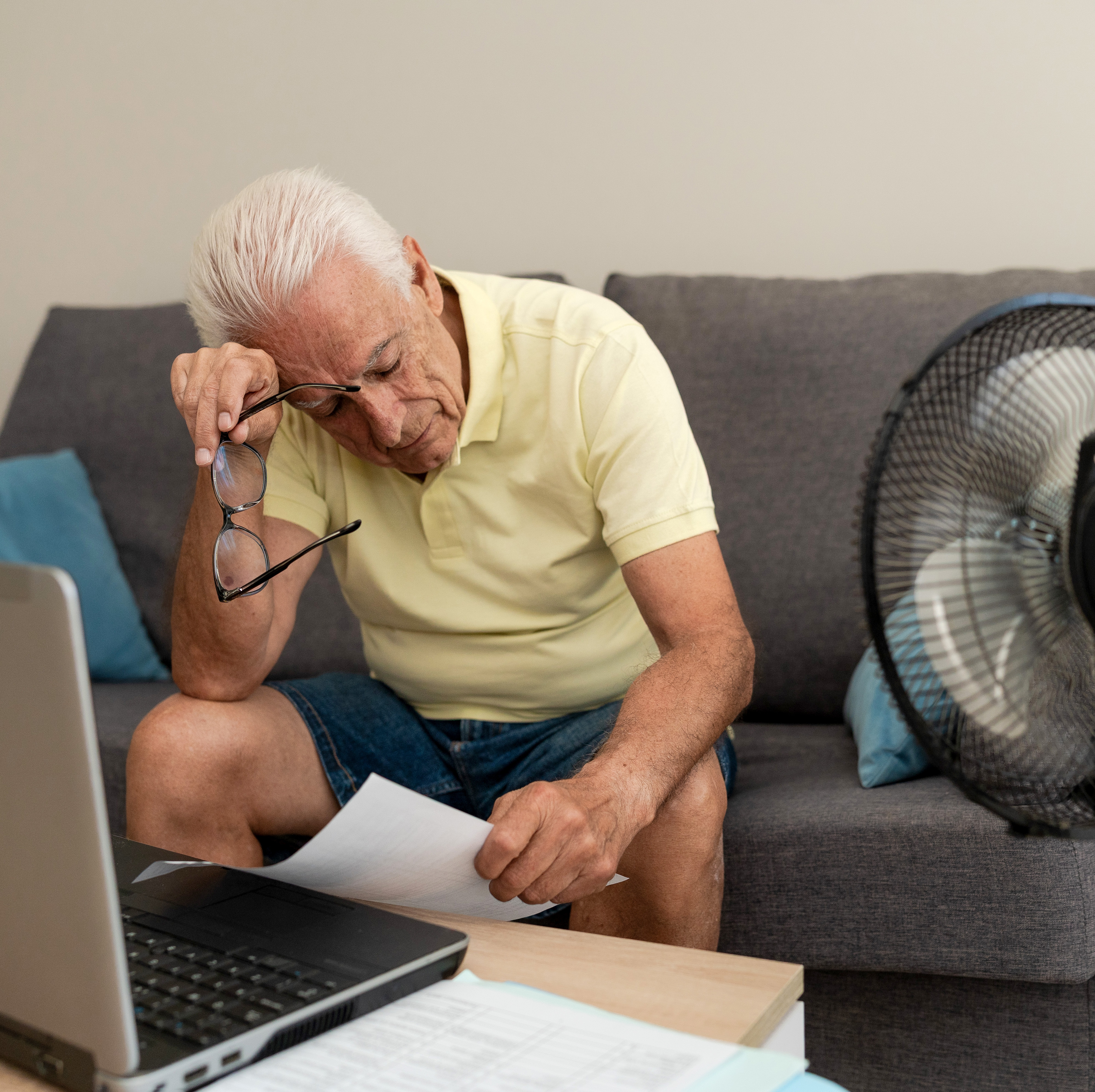 An older man sits on a couch with a fan close by blowing on him. A laptop sits on the coffee table in front of him and he wipes his brow.
