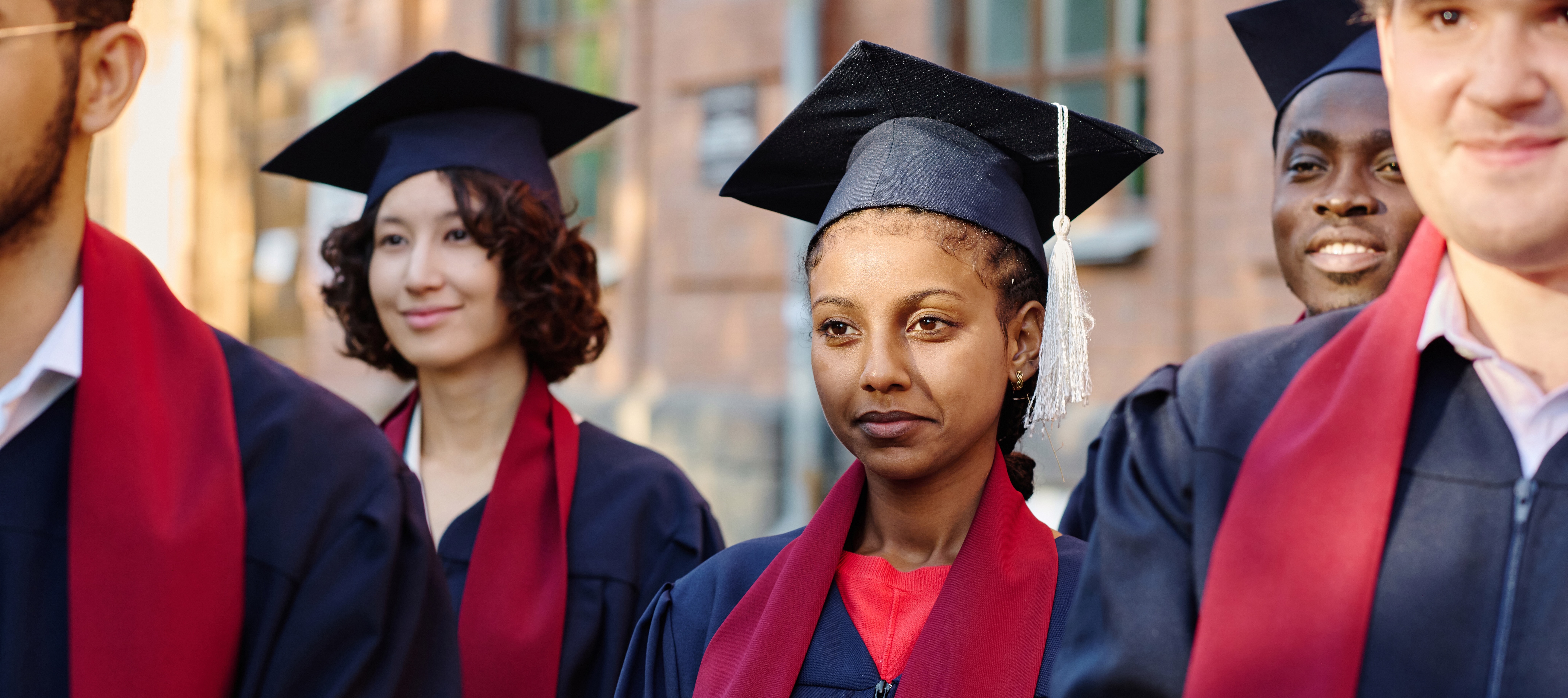 A close up of a group of young people stand together in graduation attire, wearing dark blue gowns and caps with red stoles.