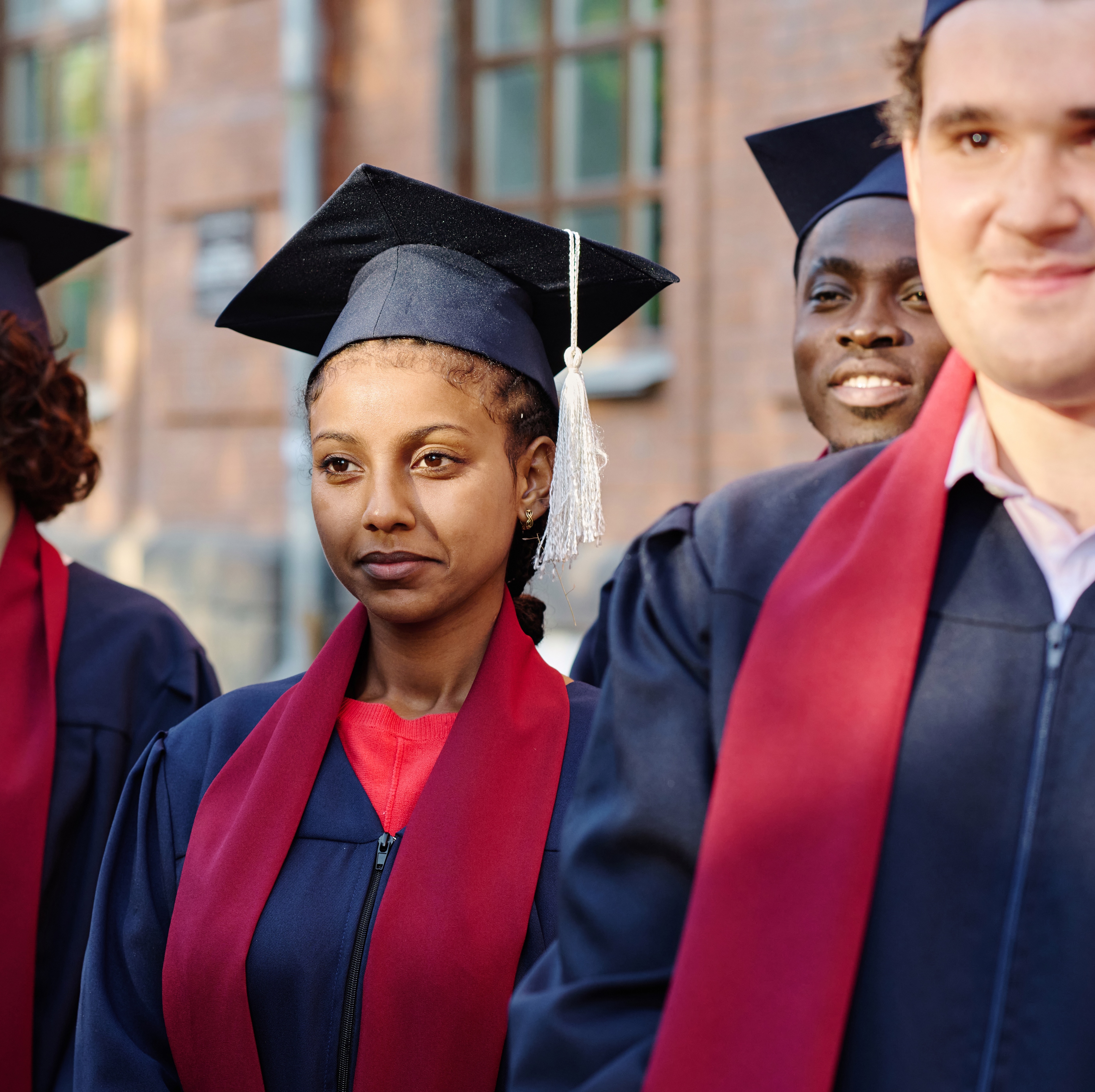 A close up of a group of young people stand together in graduation attire, wearing dark blue gowns and caps with red stoles.