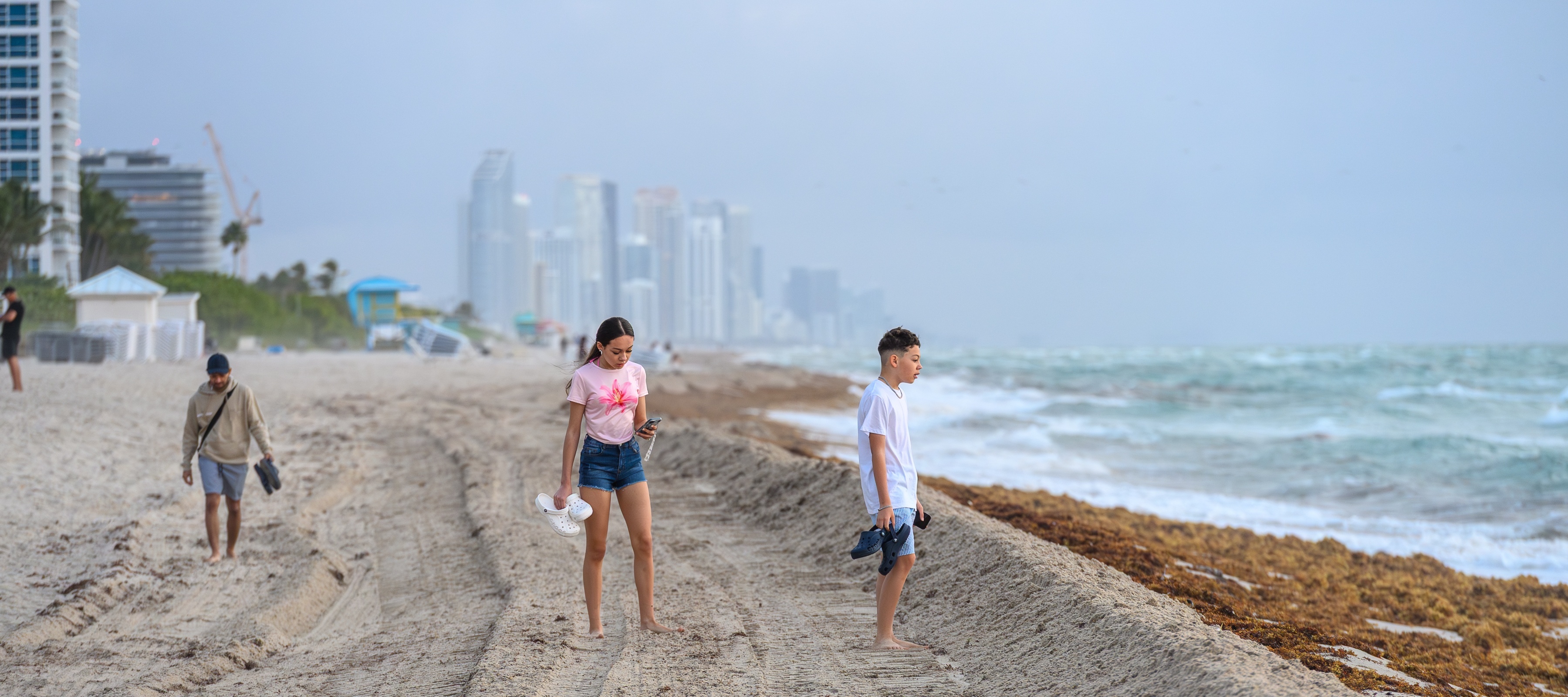 Young people walking on a sandy shore with tire tracks seaweed and distant city buildings in Miami Florida USA.
