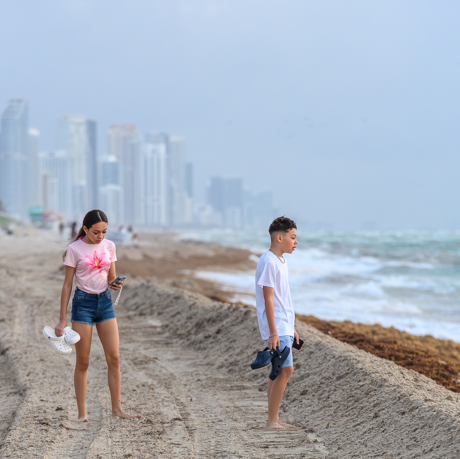Young people walking on a sandy shore with tire tracks seaweed and distant city buildings in Miami Florida USA.