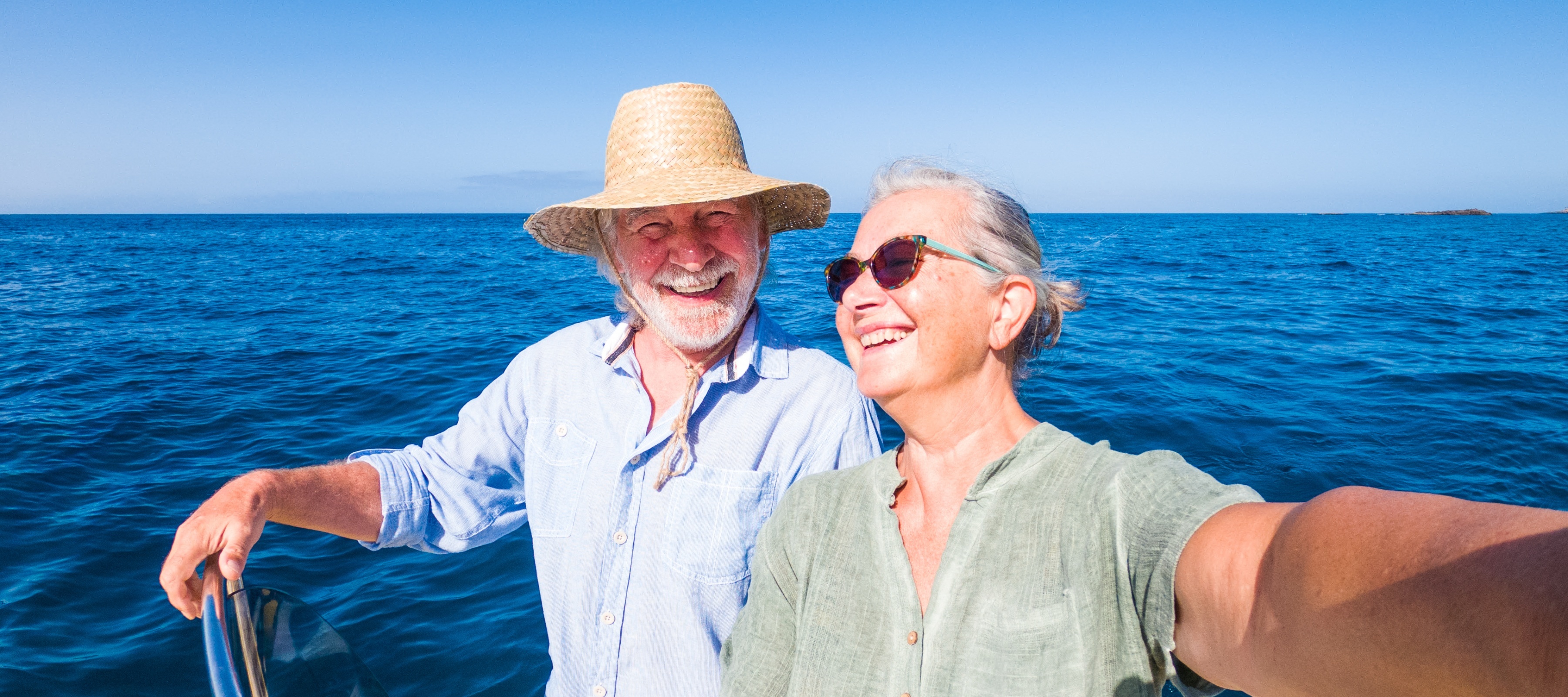 Older couple smiling, taking a selfie on a boat in front of shimmering blue waters.