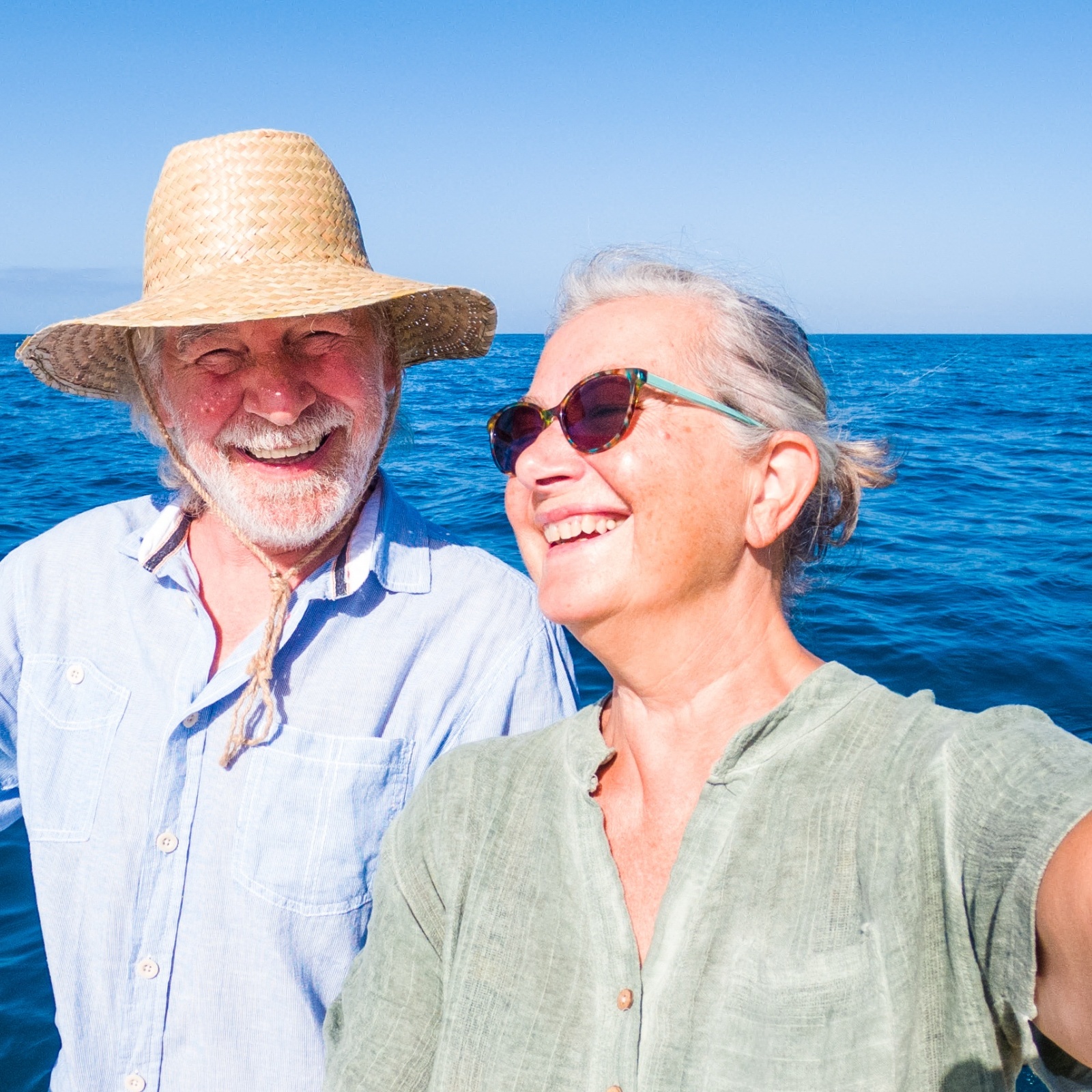 Older couple smiling, taking a selfie on a boat in front of shimmering blue waters.